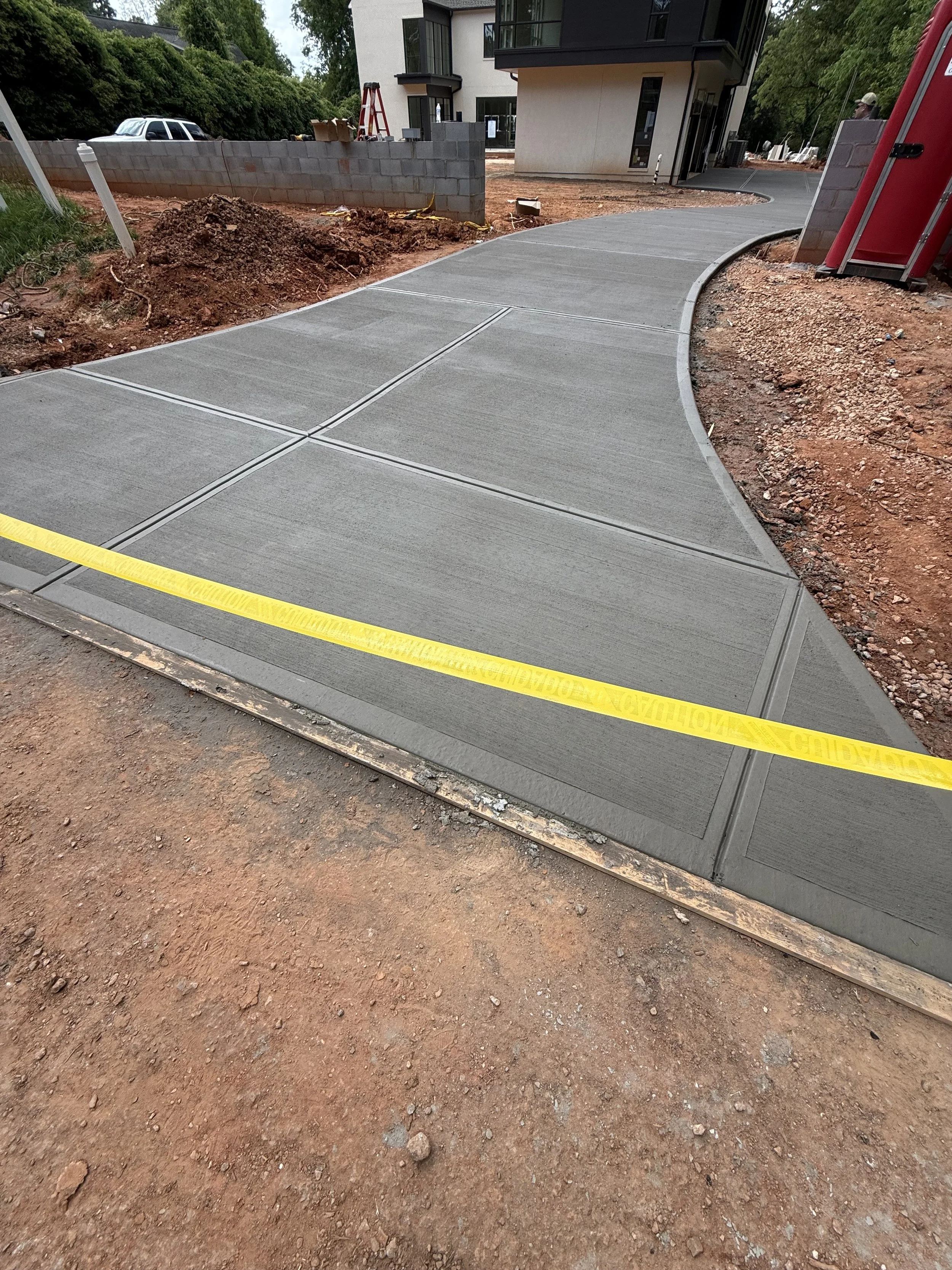 Newly poured concrete sidewalk with expansion joints, curb, yellow caution tape, and construction site dirt and debris, with a building and parked cars in the background.