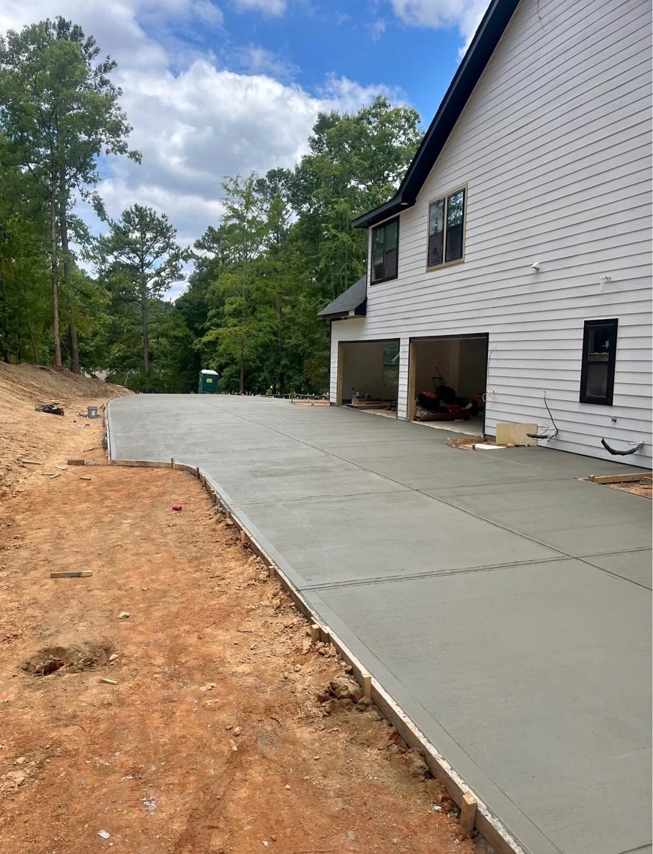 Newly poured concrete driveway next to a white house under construction, with a backdrop of trees and a partly cloudy sky.