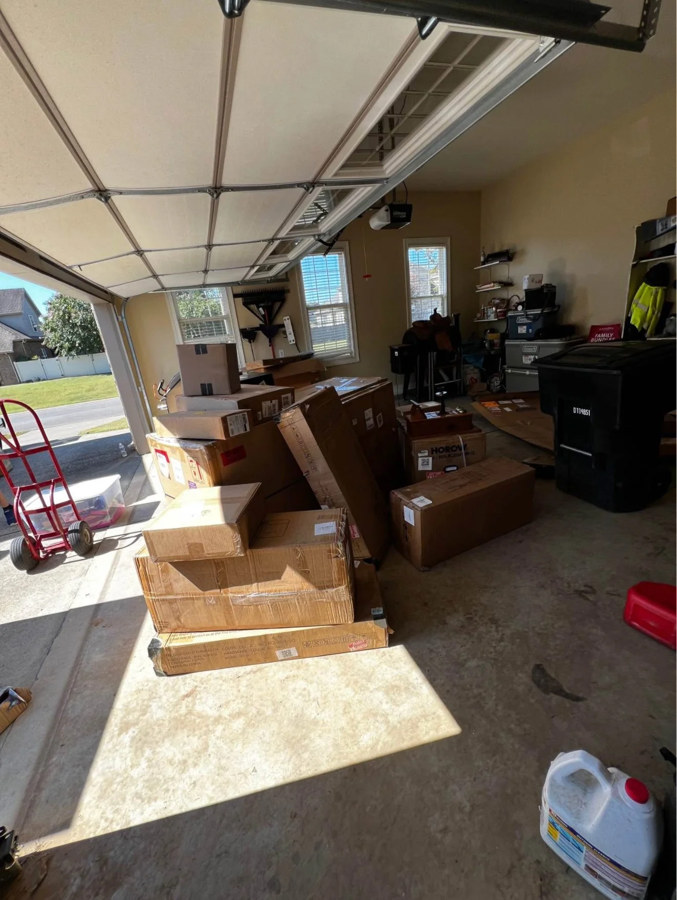 Garage with several stacked cardboard boxes, a red hand truck, a black storage bin, and various tools and supplies on shelves.