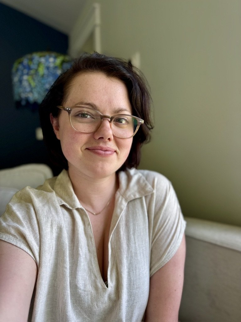 A woman with short dark hair and glasses smiling at the camera, sitting indoors in a well-lit room.
