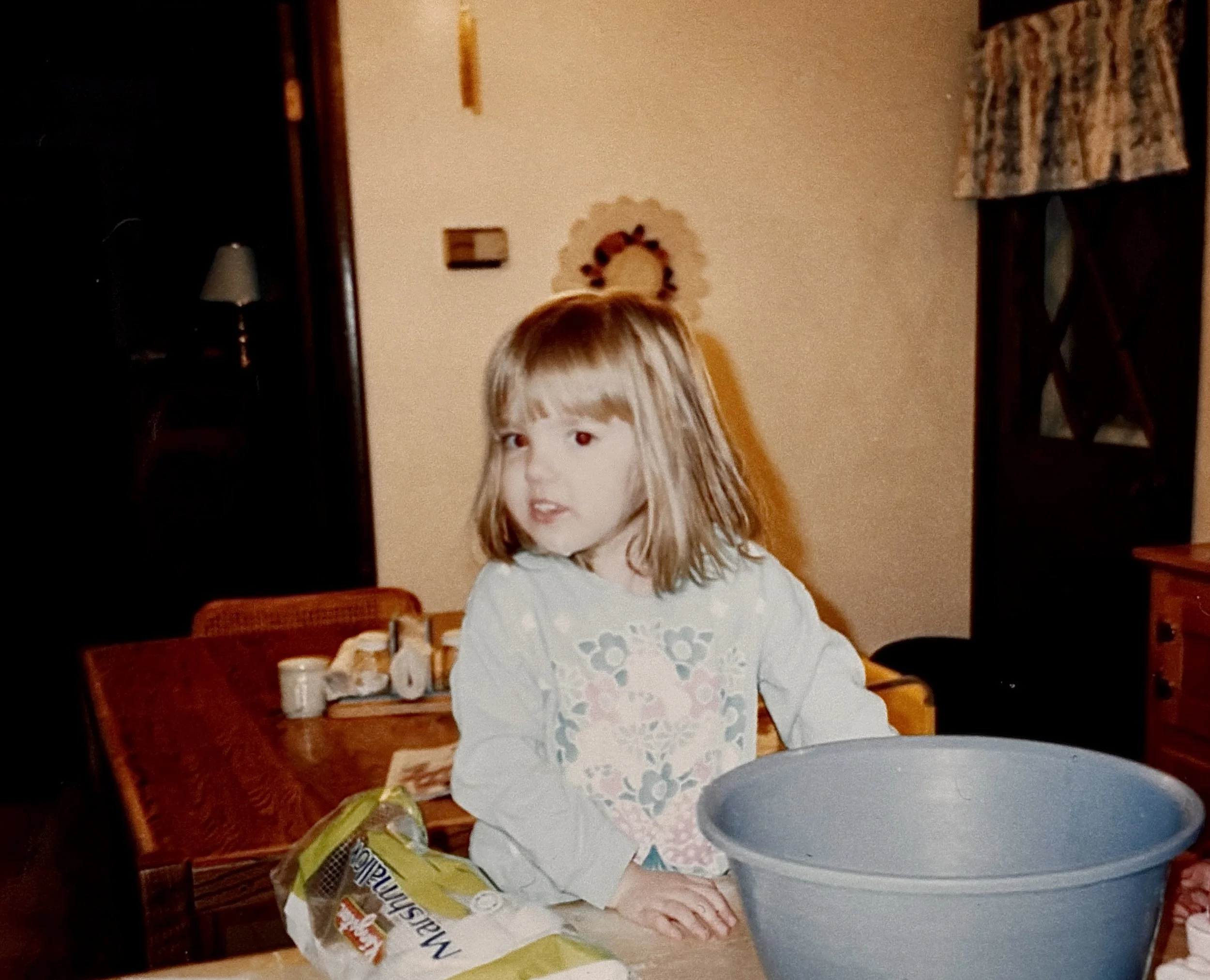 A young girl with shoulder-length blonde hair and bangs standing at a table in a kitchen or dining room, with a blue mixing bowl in front of her and a bag of bread on the table.