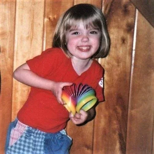 A young girl smiling and holding a colorful fidget toy, standing in front of a wooden wall.