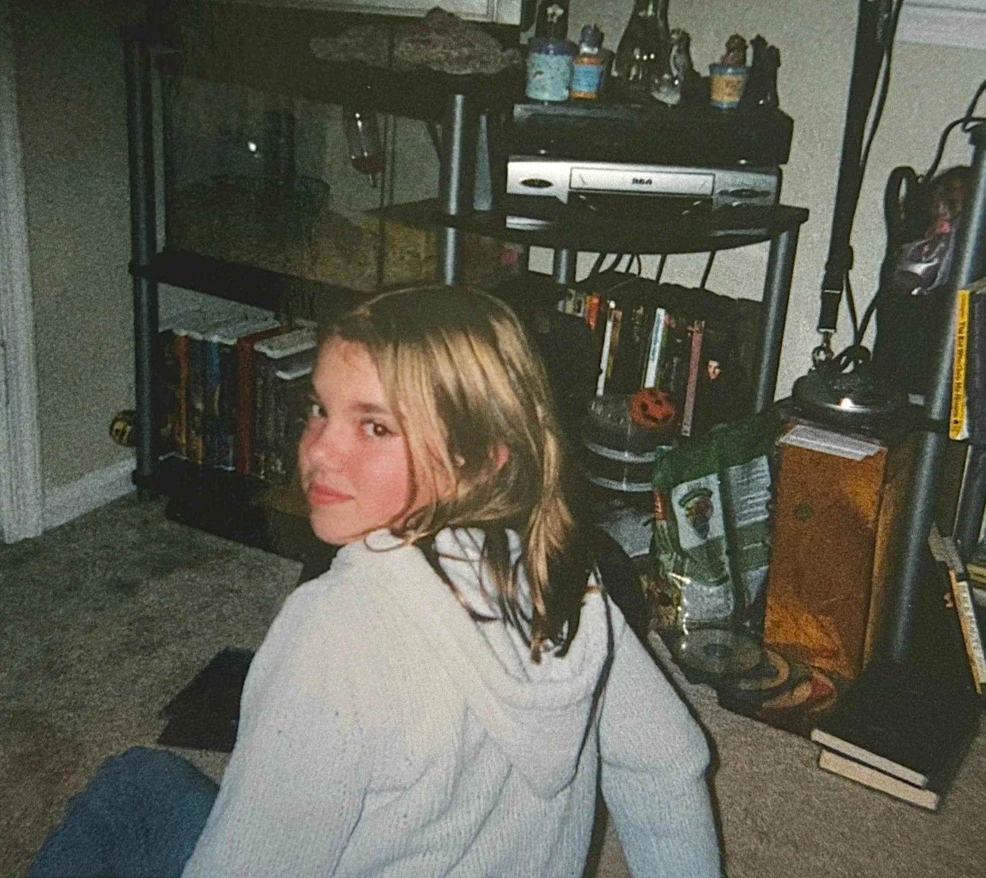 A young woman with shoulder-length light brown hair, sitting on a carpeted floor, looking over her shoulder toward the camera. She is wearing a light-colored sweater.