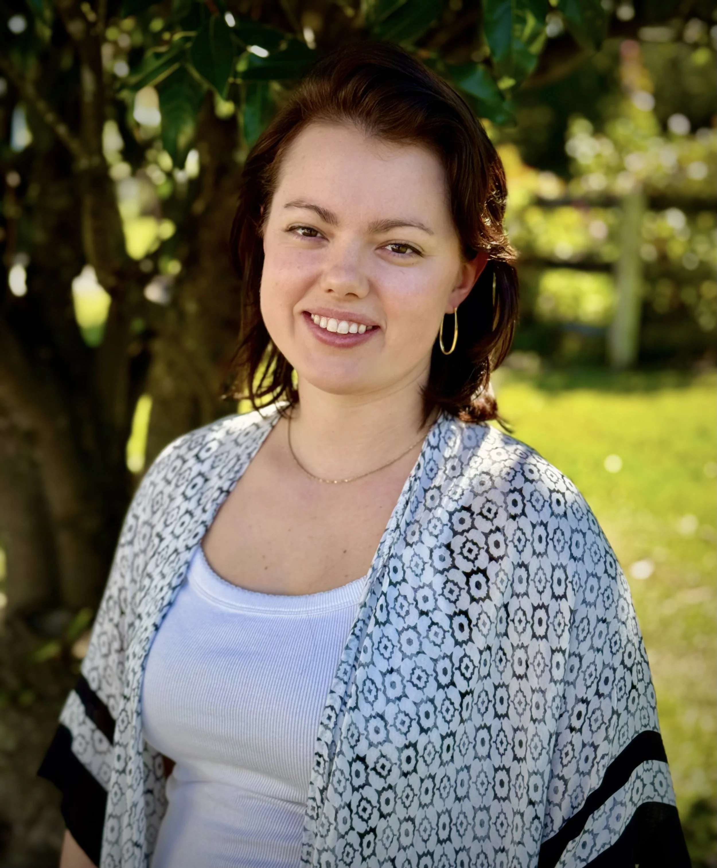 A young woman with short brown hair smiling outdoors in front of green trees and grass, wearing a white top, patterned cover-up, hoop earrings, and a delicate necklace.