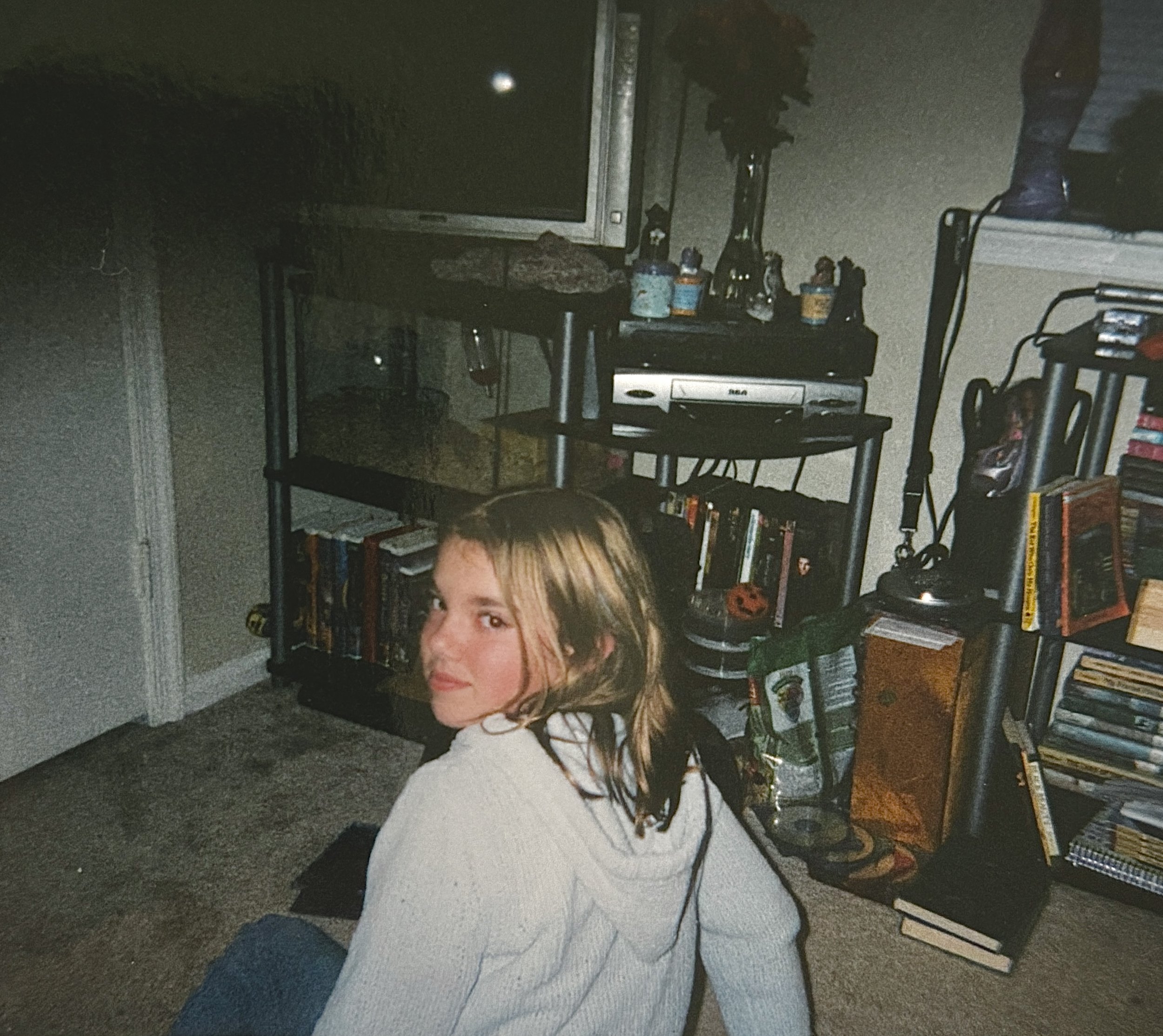 A young woman with blonde hair sitting on the floor inside a room, looking at the camera, with bookshelves and a television behind her.