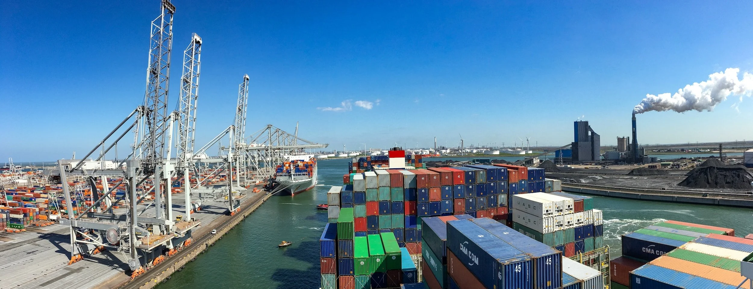 Container port with stacked shipping containers, cranes, large cargo ship, and industrial buildings emitting smoke under a blue sky.