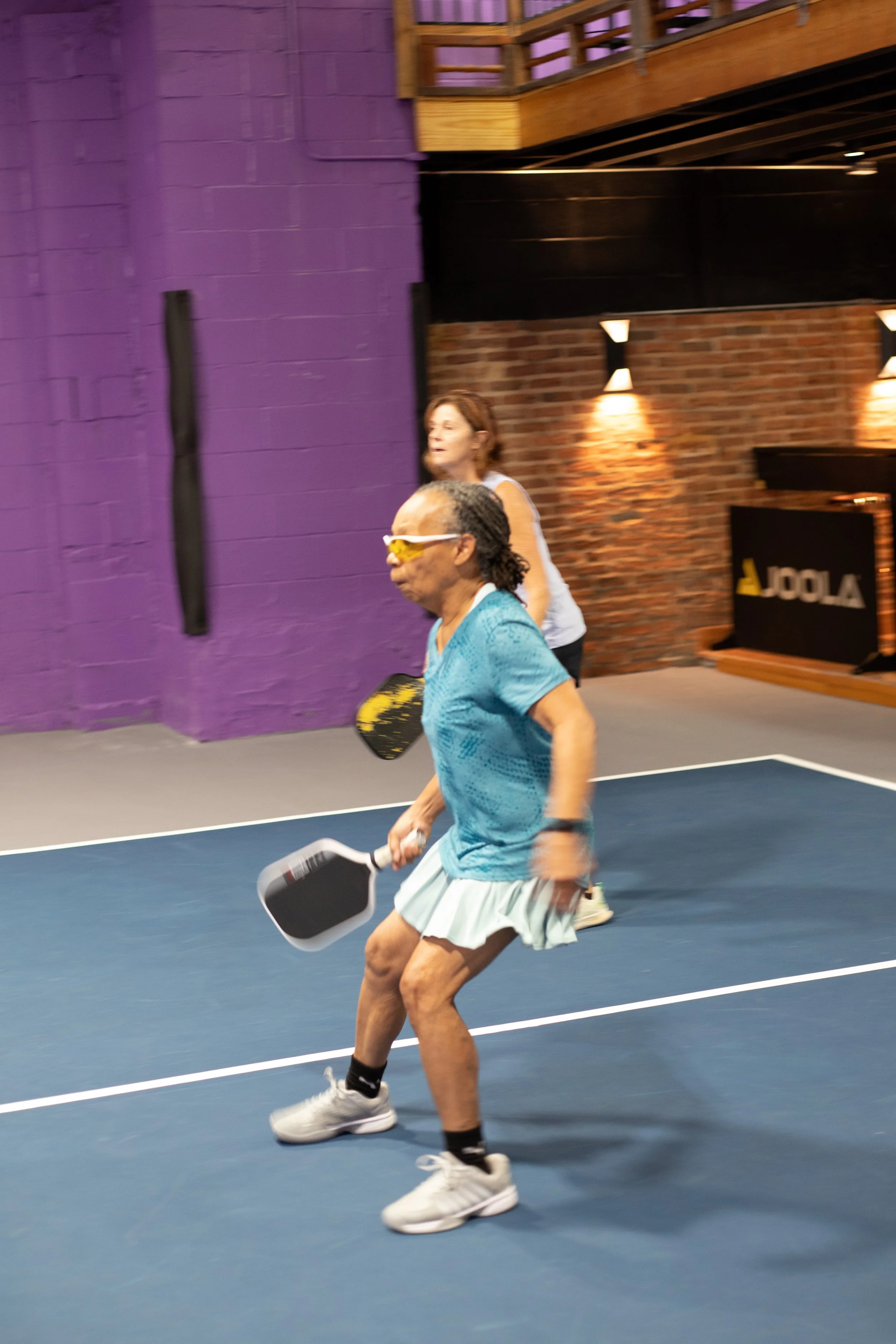 A man with curly dark hair, glasses, and a beard, wearing a black sweater, sitting indoors with a plant in the background.