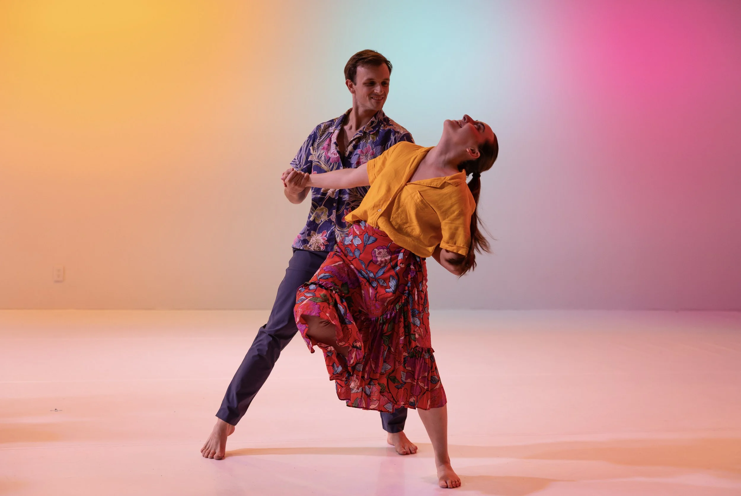 Two dancers perform a partnered dip under colorful stage lighting, bodies angled in a lively, theatrical pose.