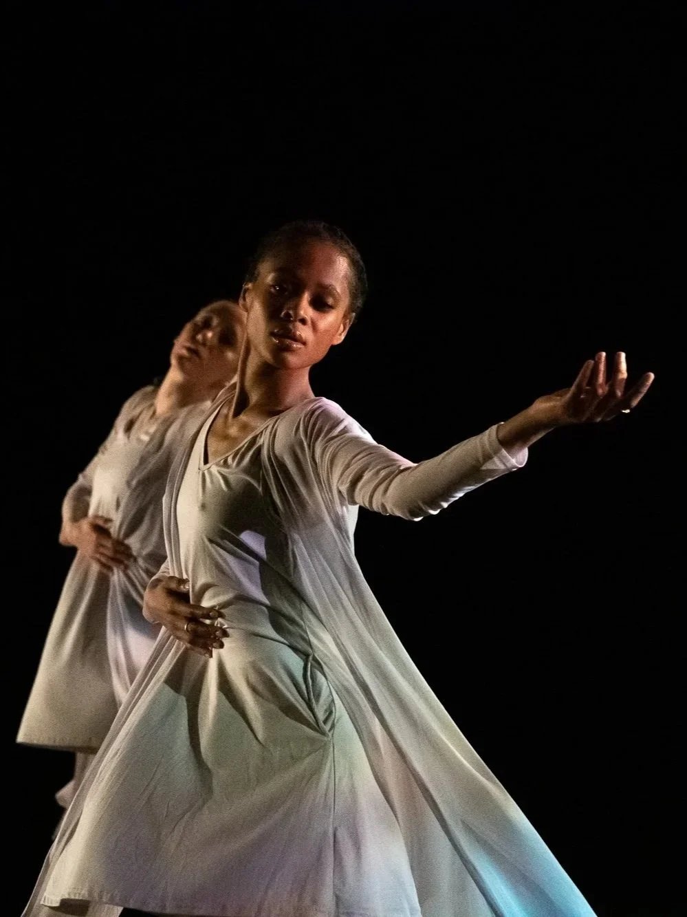 Two dancers in light costumes move in close proximity, one extending an arm outward against a dark stage.