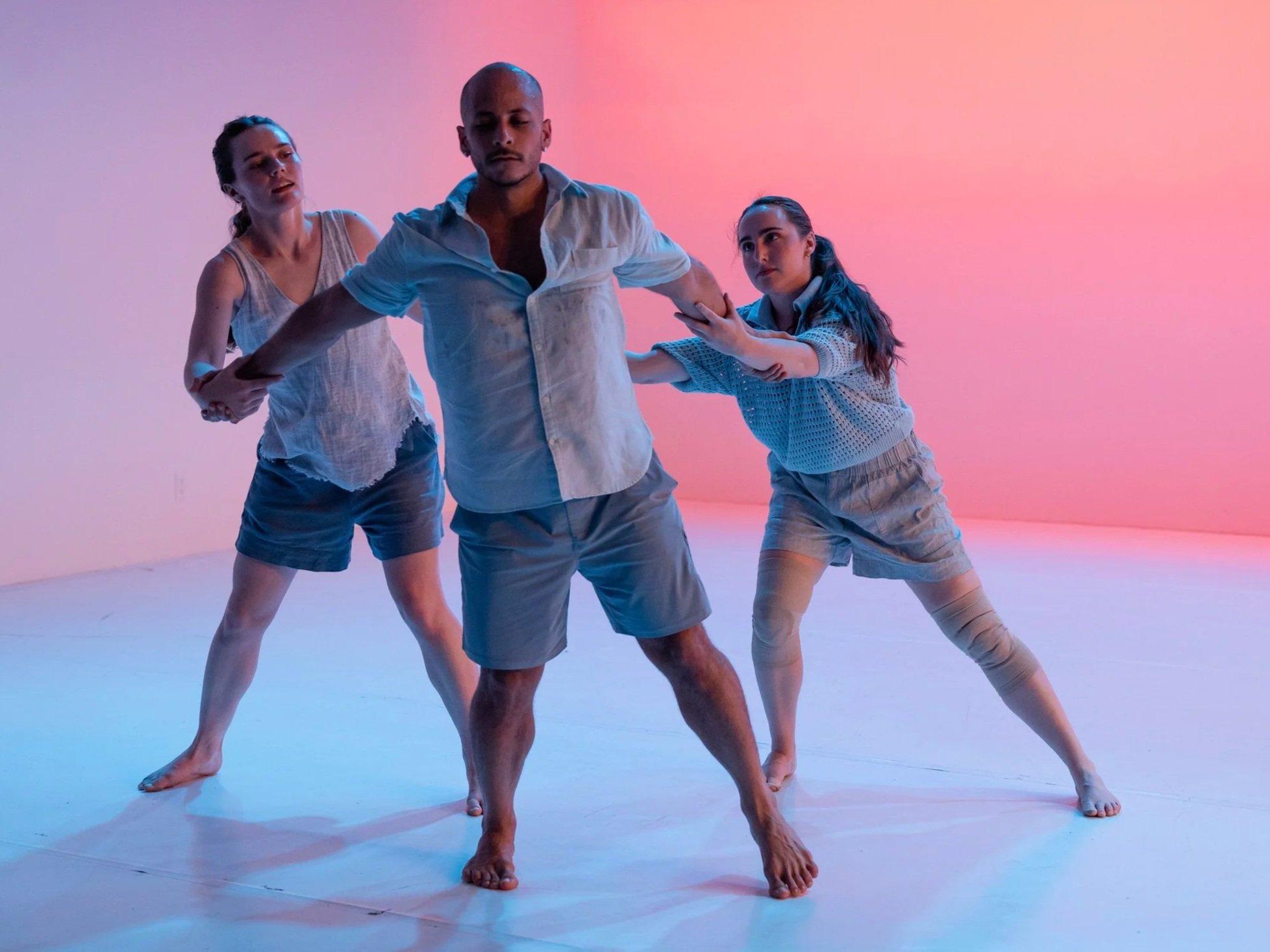Three contemporary dancers move together in a grounded formation, two supporting a central dancer by the arms, as they step forward across a white stage lit with soft pink and blue gradients.