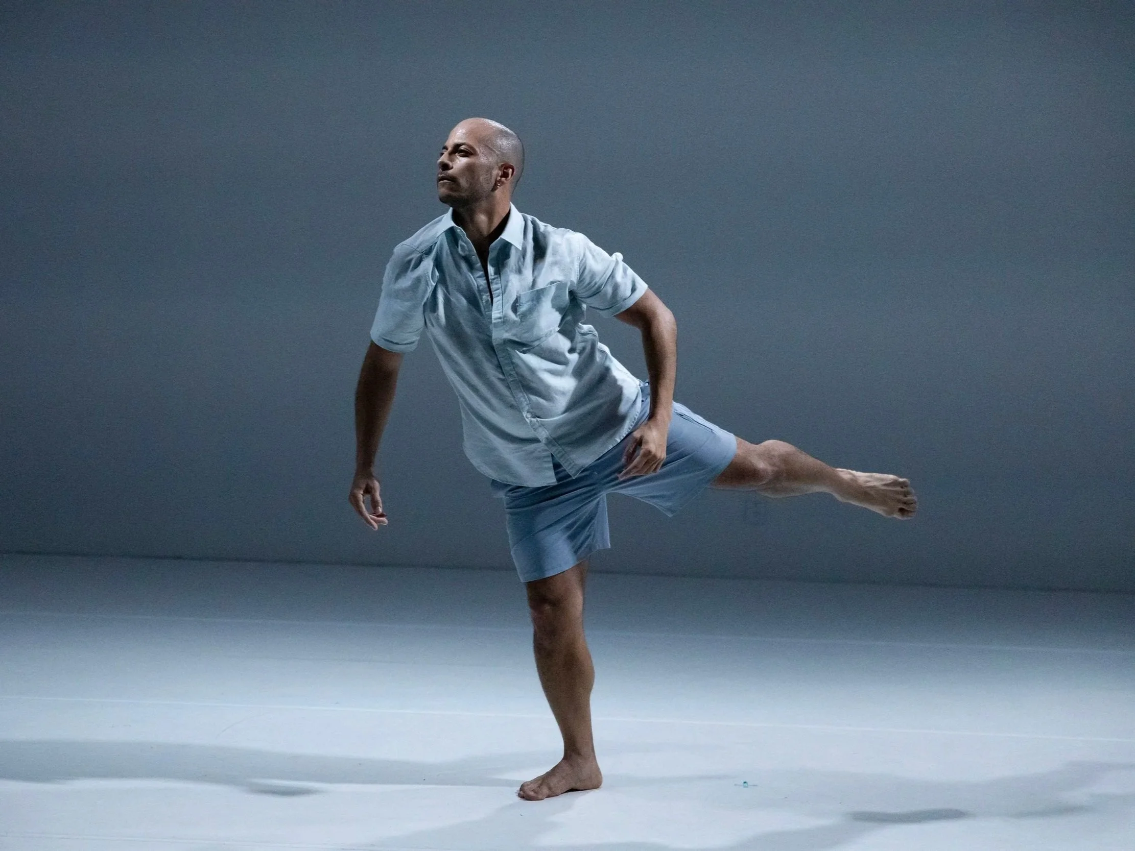 A contemporary dancer balances on one leg with the other extended behind, wearing a light blue shirt and shorts, moving calmly across a minimalist stage under cool gray lighting.