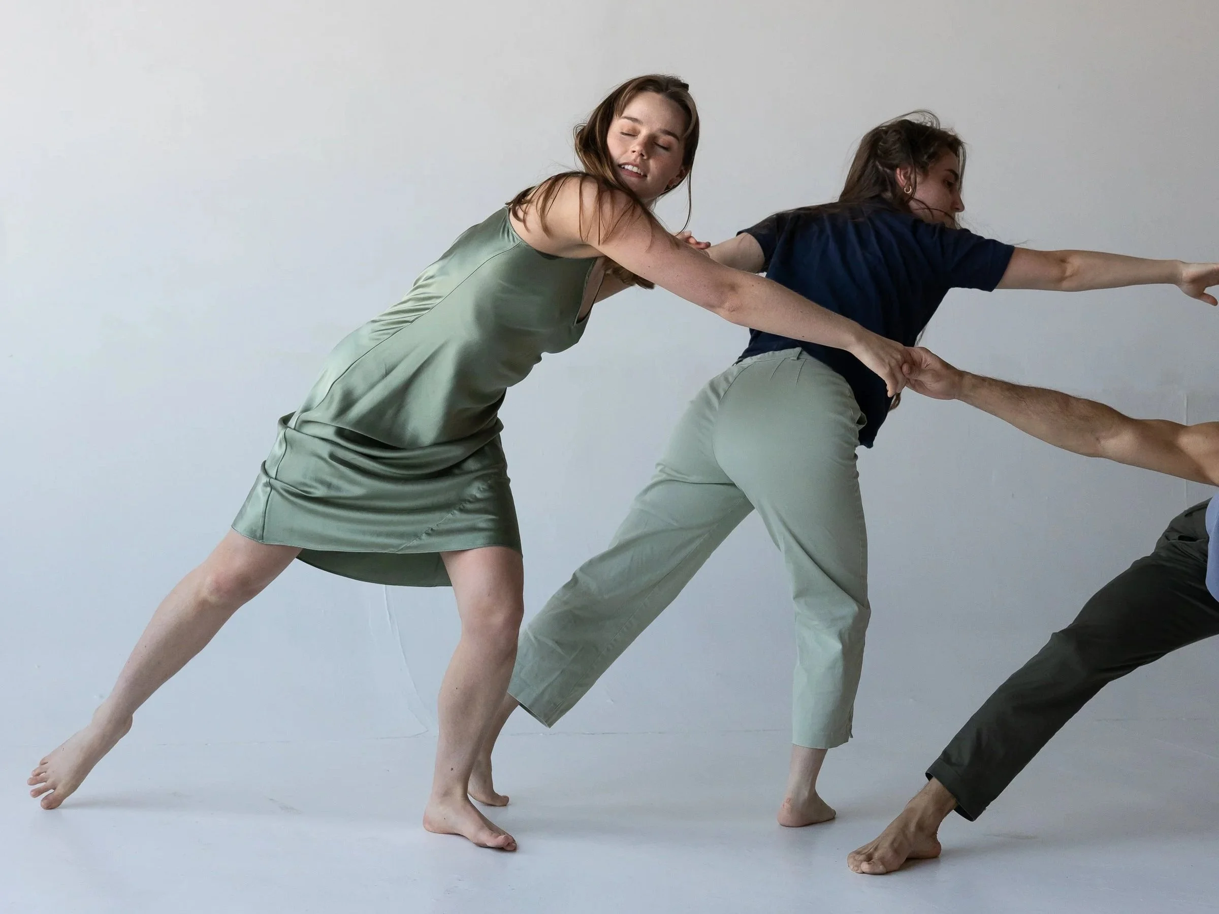 Three dancers move diagonally across the floor, linked by hands and momentum, as one leans forward and another reaches outward, suggesting motion, pull, and transition.