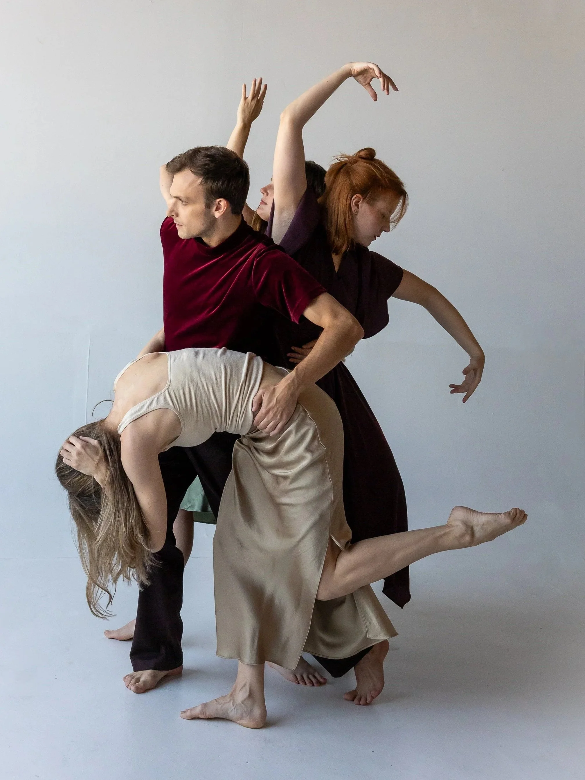 Dancers form a vertical, intertwined composition as one dancer is bent forward and supported at the waist, while others extend arms overhead and behind, shaping a layered, asymmetrical structure.