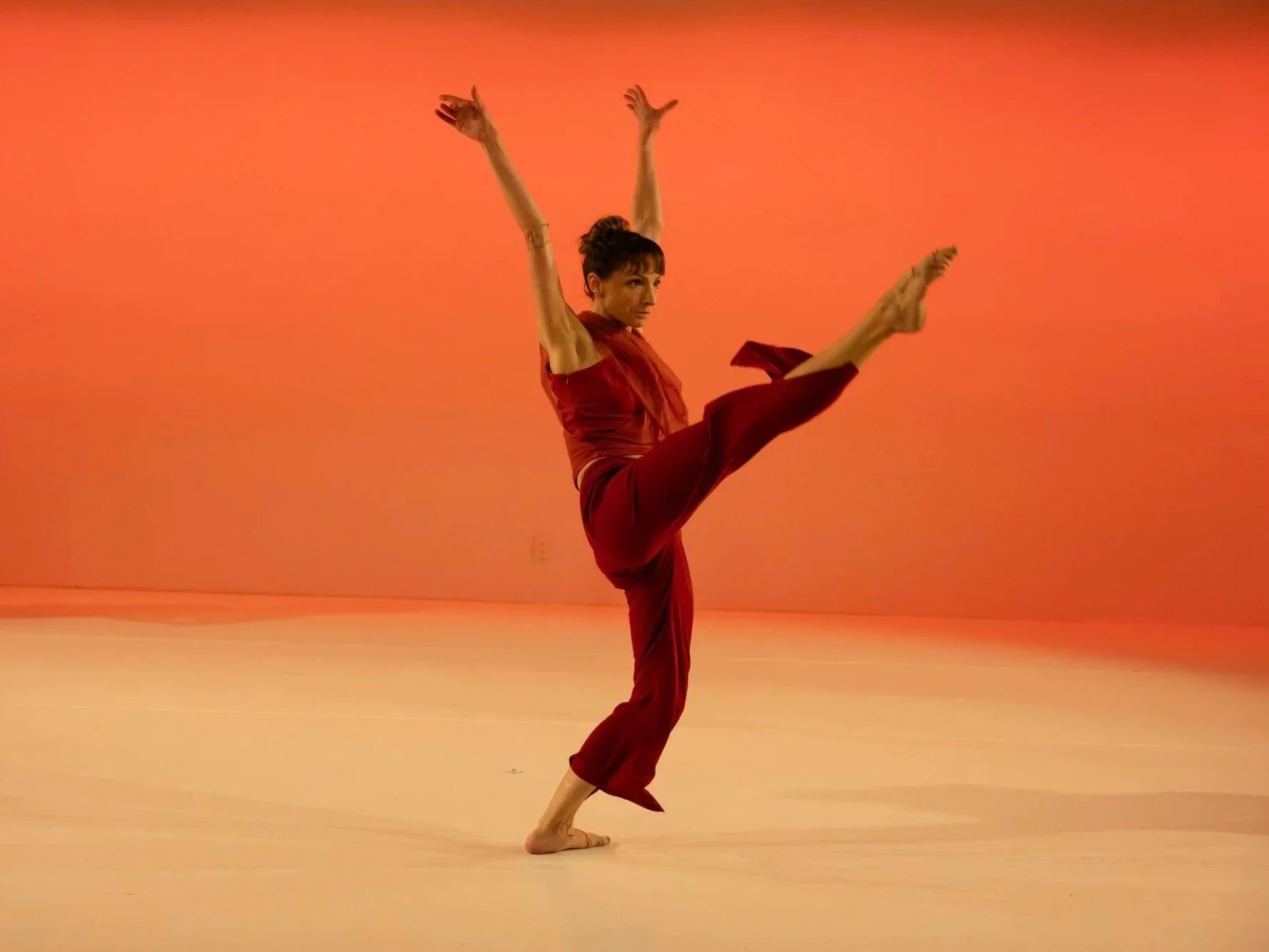 A solo dancer in red lifts one leg high with arms raised, performing against a warm orange backdrop.