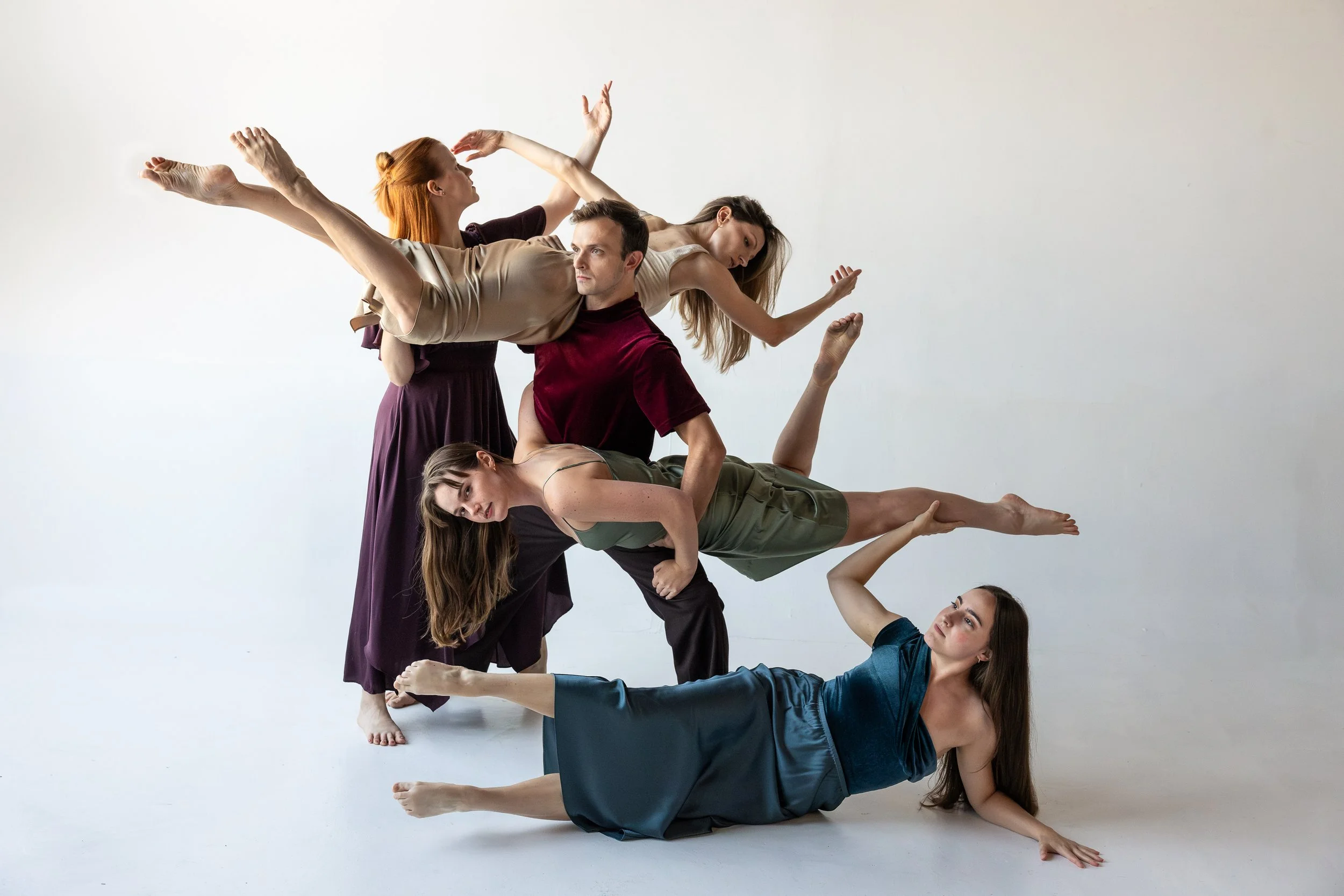 A male dancer anchors a complex group lift as multiple dancers extend horizontally in opposing directions, forming a dynamic tableau of suspended bodies and counterbalanced weight.