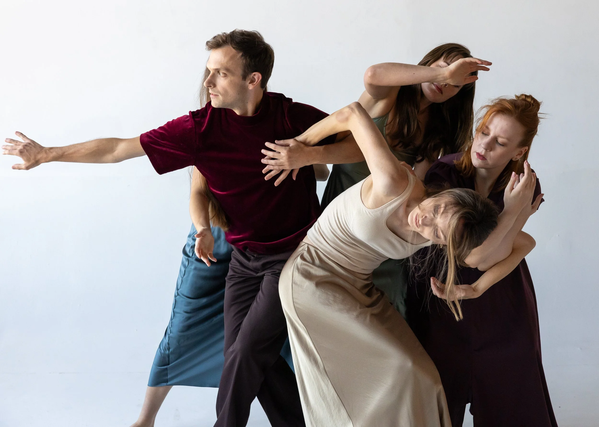 A group of contemporary dancers interlock in a sculptural formation, with one dancer reaching outward while others fold and curve around them, creating a sense of tension and collective support.