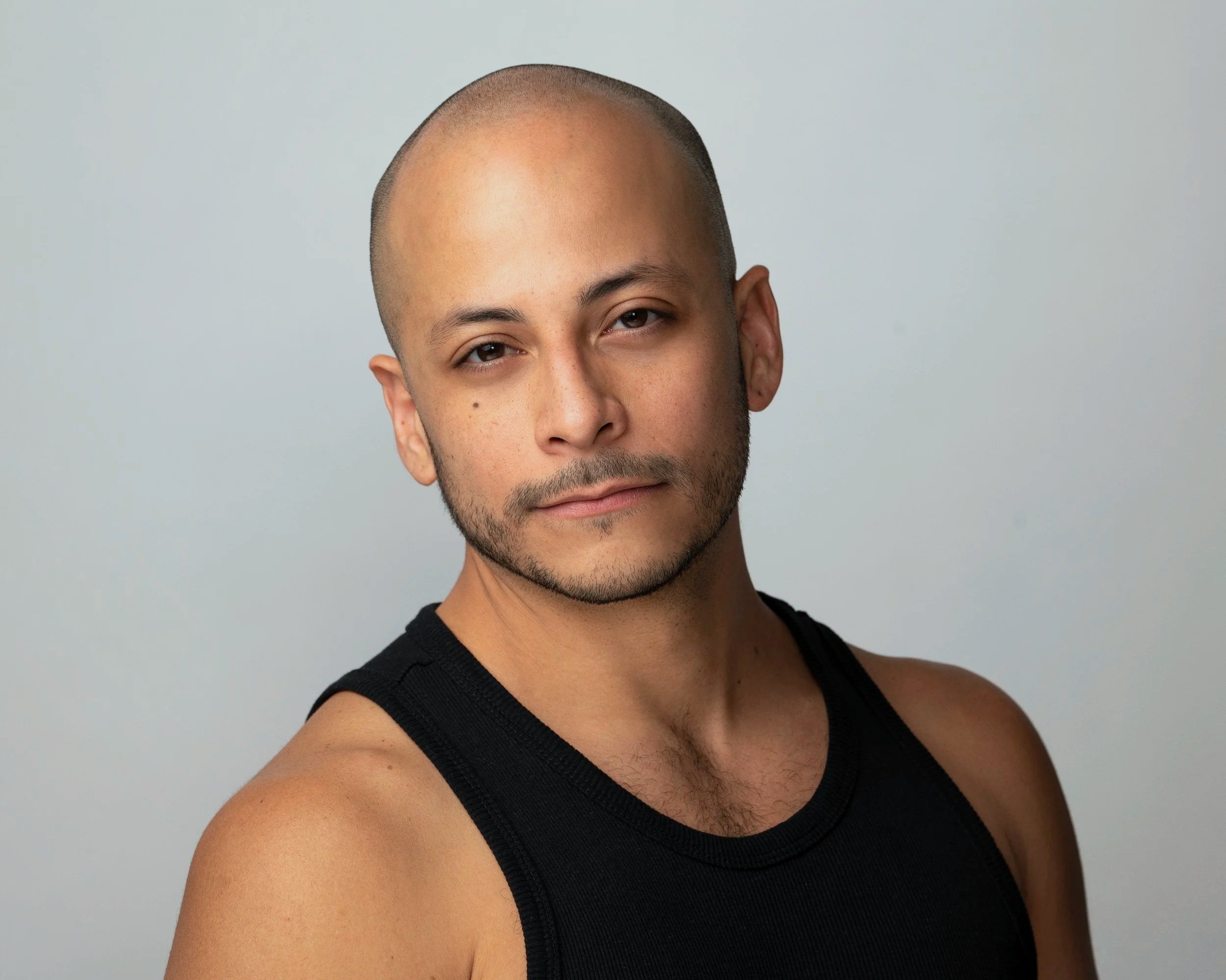 A man with a shaved head and trimmed beard looks calmly toward the camera against a light background.