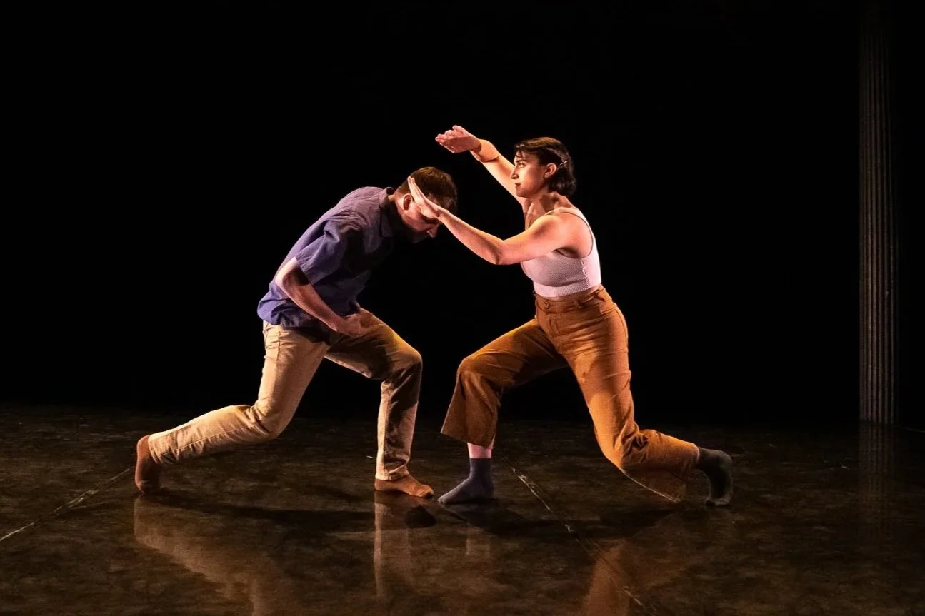 Two dancers face each other in a grounded duet, lunging and reaching under dramatic stage lighting.