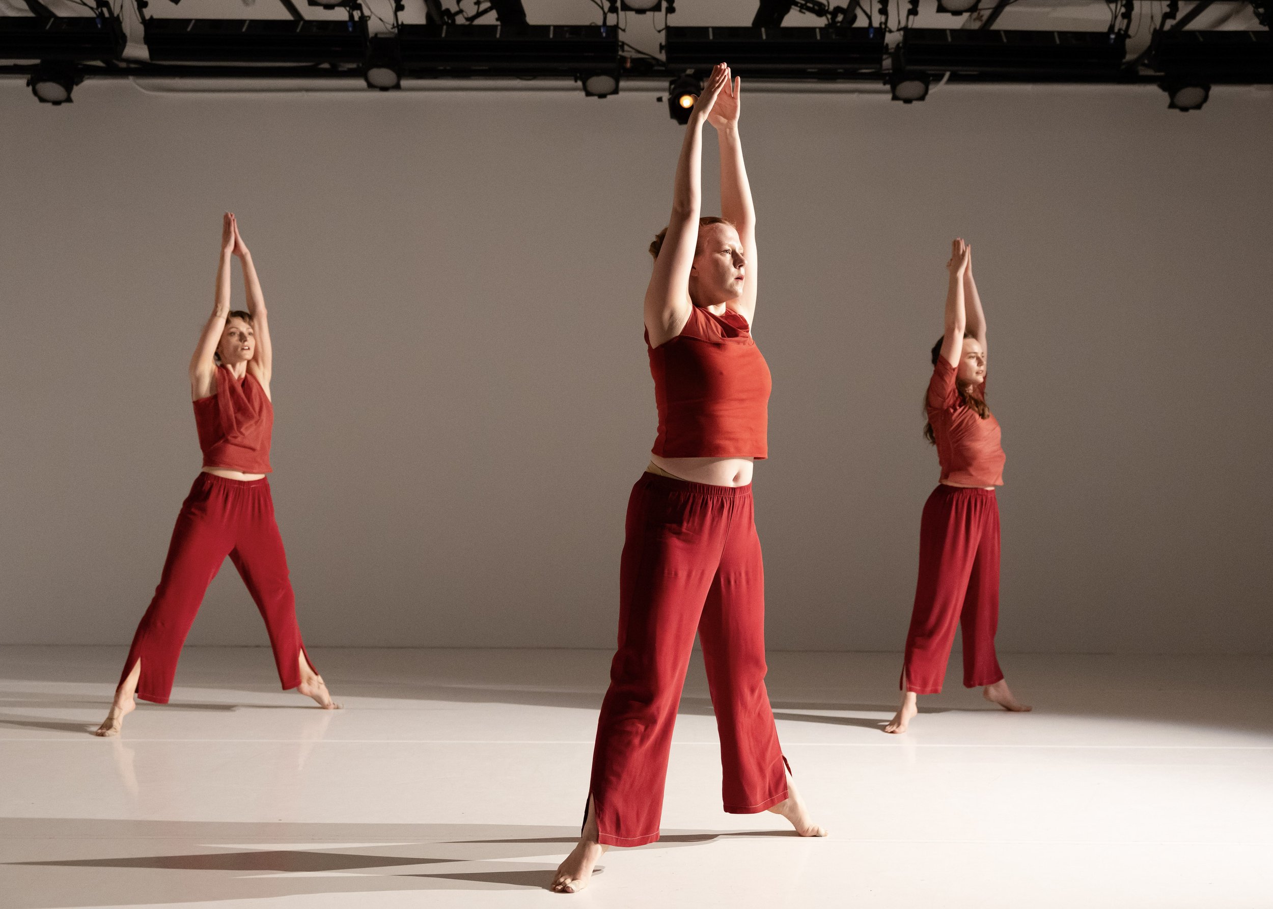 Three dancers in red costumes stand grounded with arms lifted overhead in a quiet, unified pose.