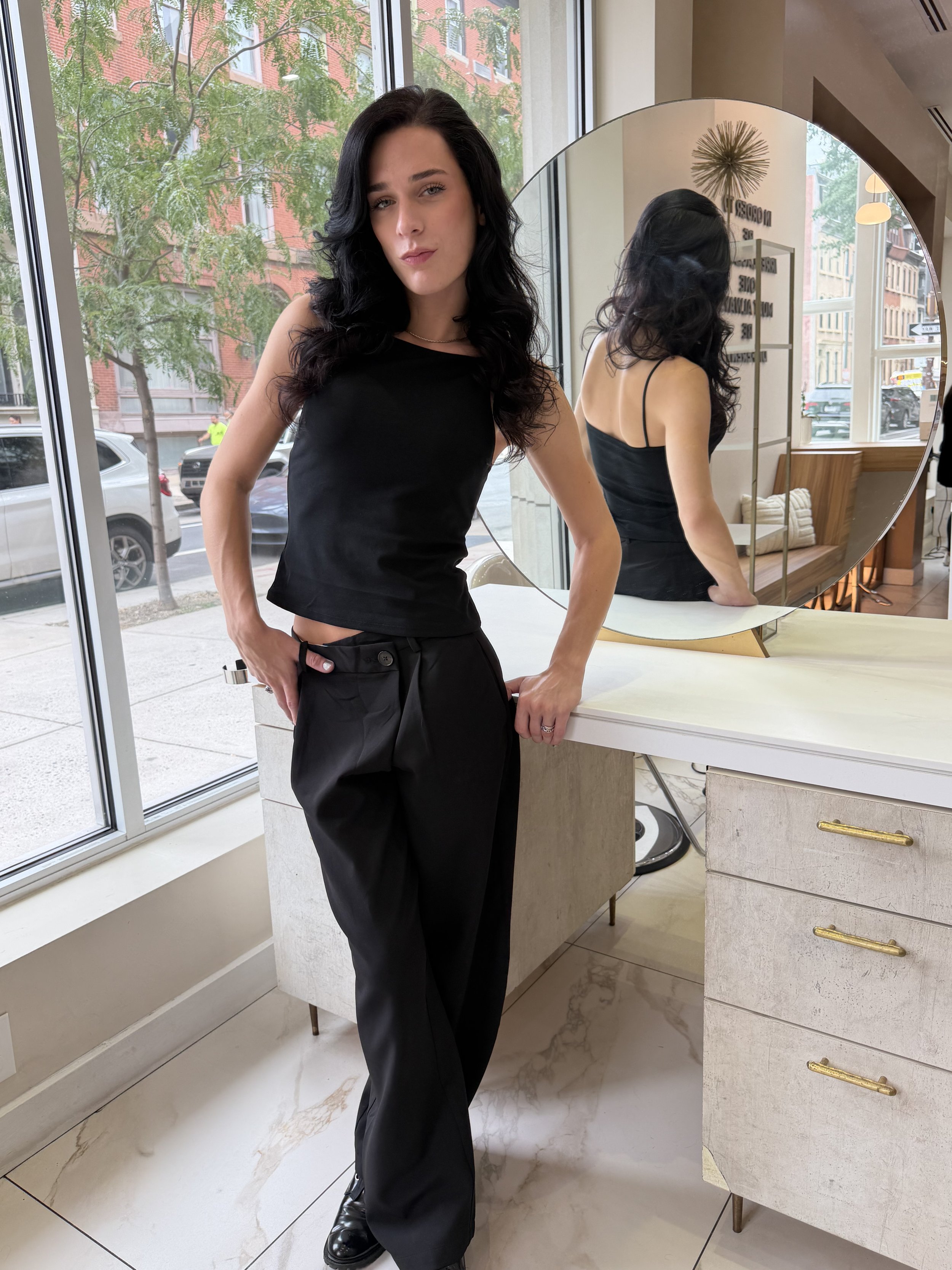 A woman with long black hair wearing a black top and black pants posing indoors in front of a large round mirror, with a window showing trees and buildings outside.