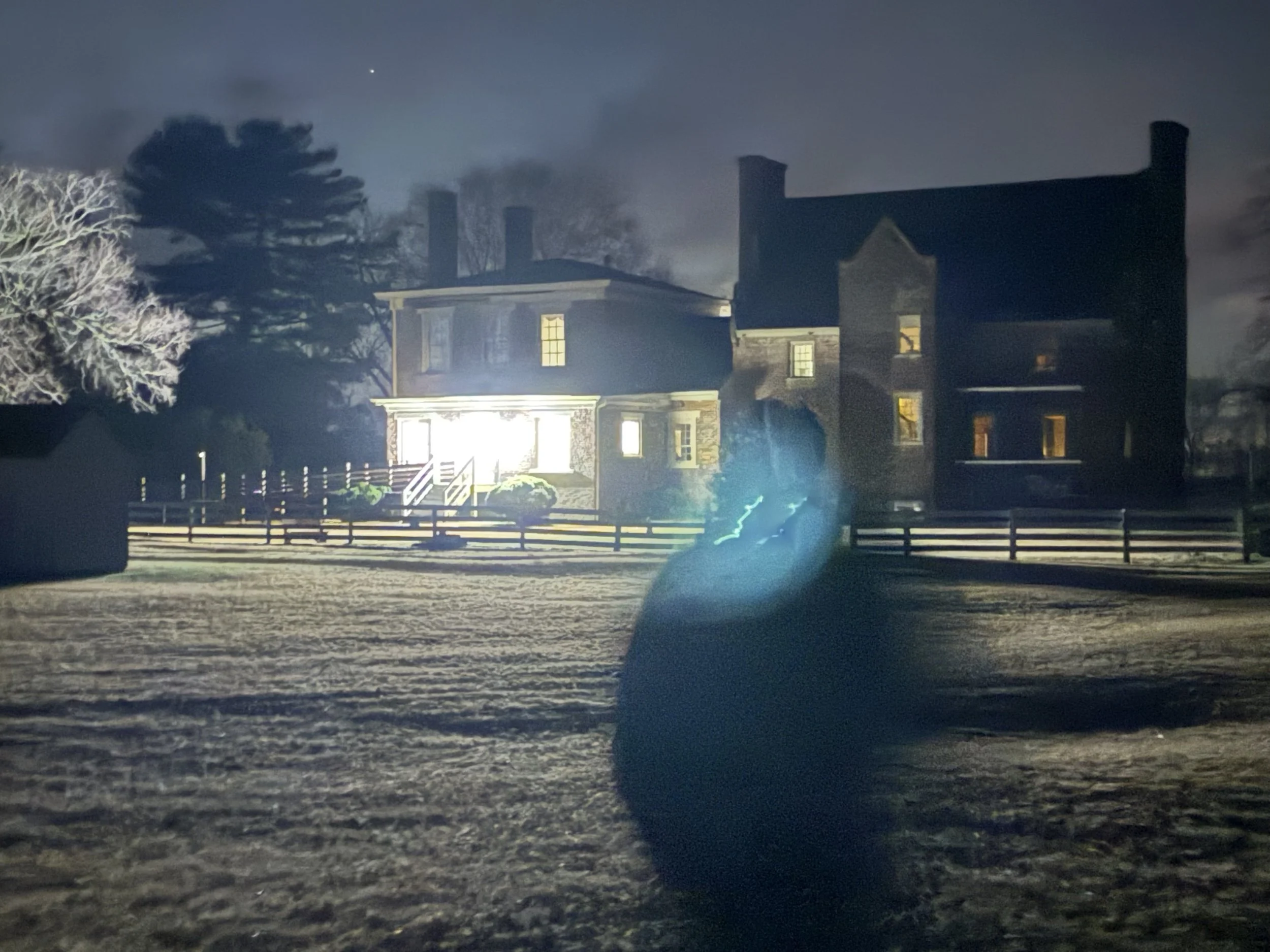 Night scene of a large, old-fashioned house illuminated from within. A tree with snow-covered branches is on the left, and a large dark silhouette of a cat is in the foreground, with a blue lightning bolt near its tail.