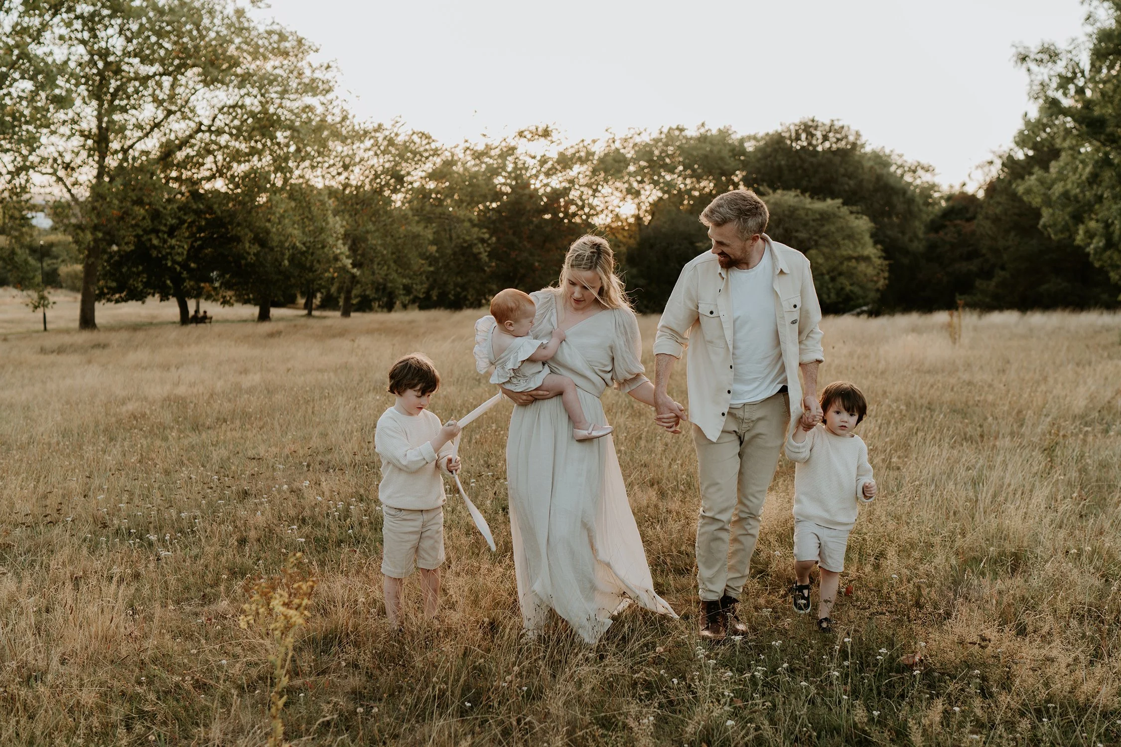 A family of five walking through a grassy field at sunset. The mother is holding a young child, and the father is holding the hand of one of their older children. The scene is natural and peaceful with trees in the background.