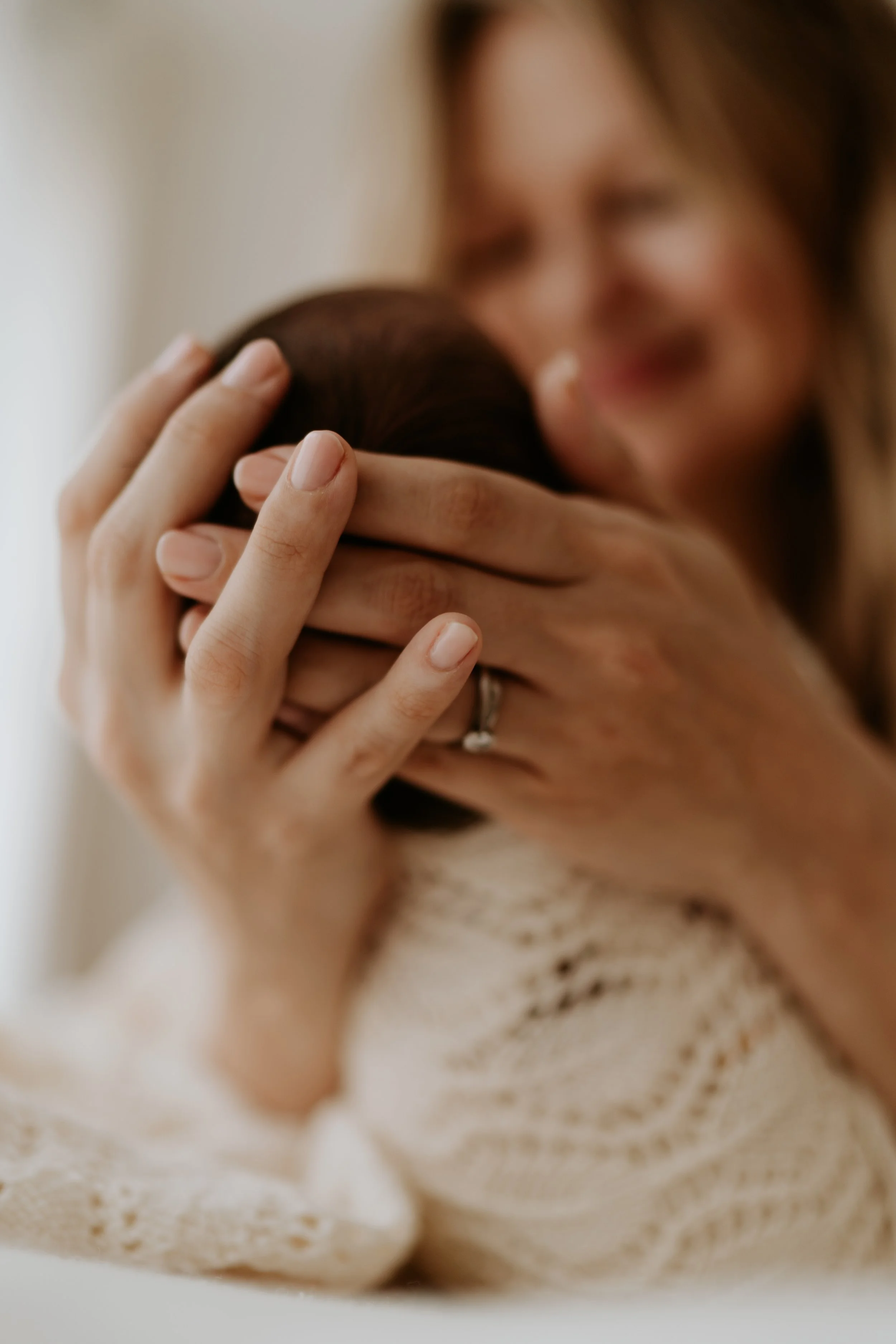 A woman holds a baby close, with her face out of focus in the background. The focus is on her hands, which gently cradle the baby's head, showing a wedding ring on her finger. The woman is wearing a white, knitted top.