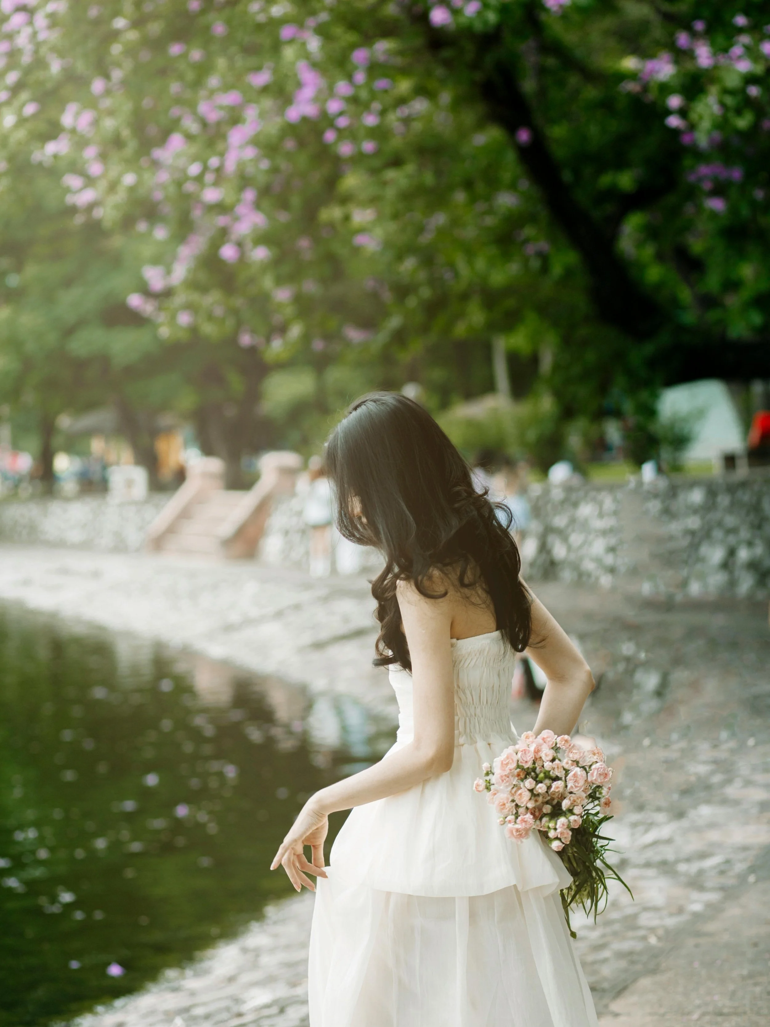 A woman in a white dress holding a bouquet of pink flowers while standing near a body of water in a park with trees and purple flowers overhead.