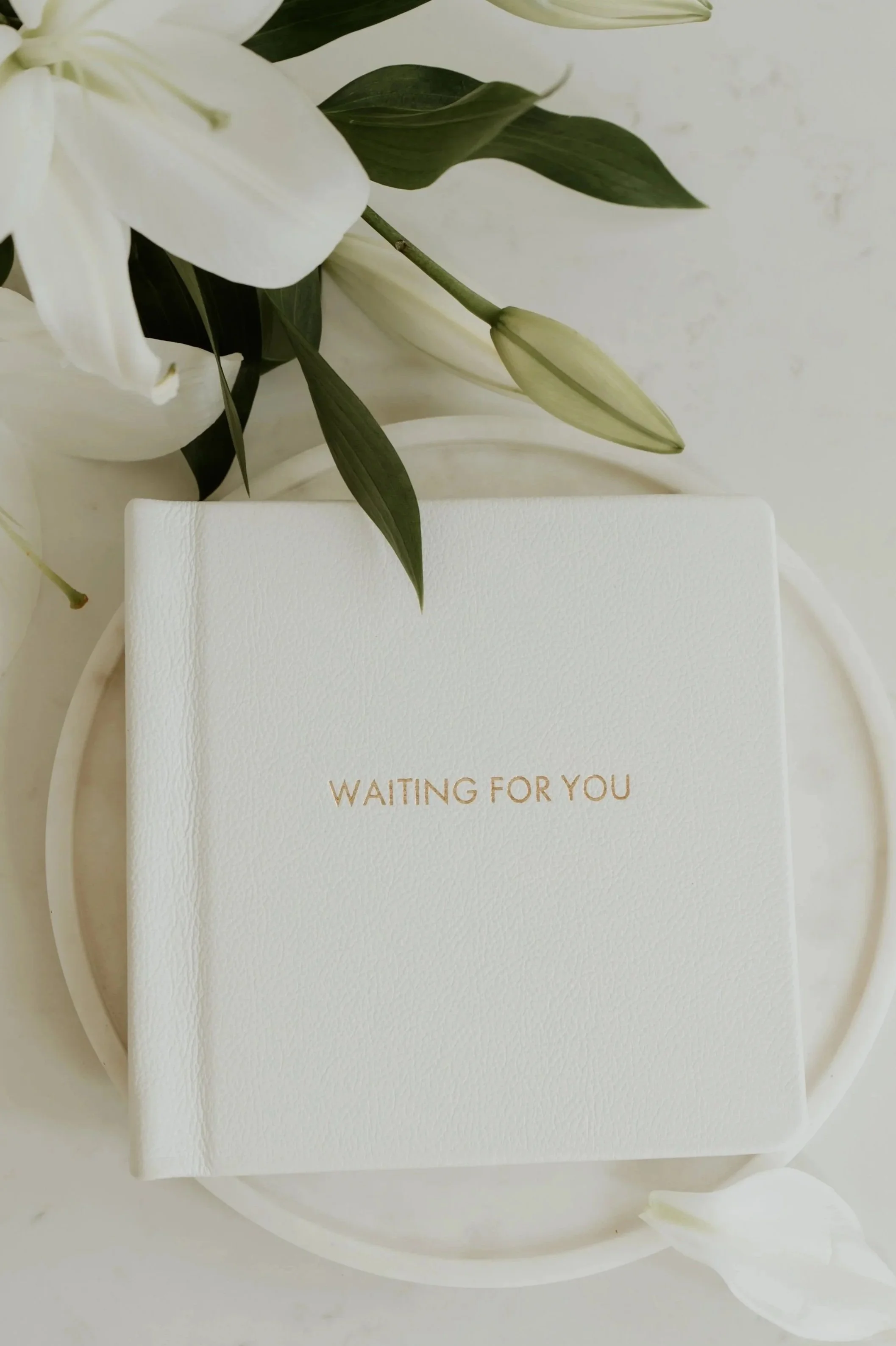 A white book with gold lettering that says 'Waiting For You' is on a white tray surrounded by white flowers and green leaves.