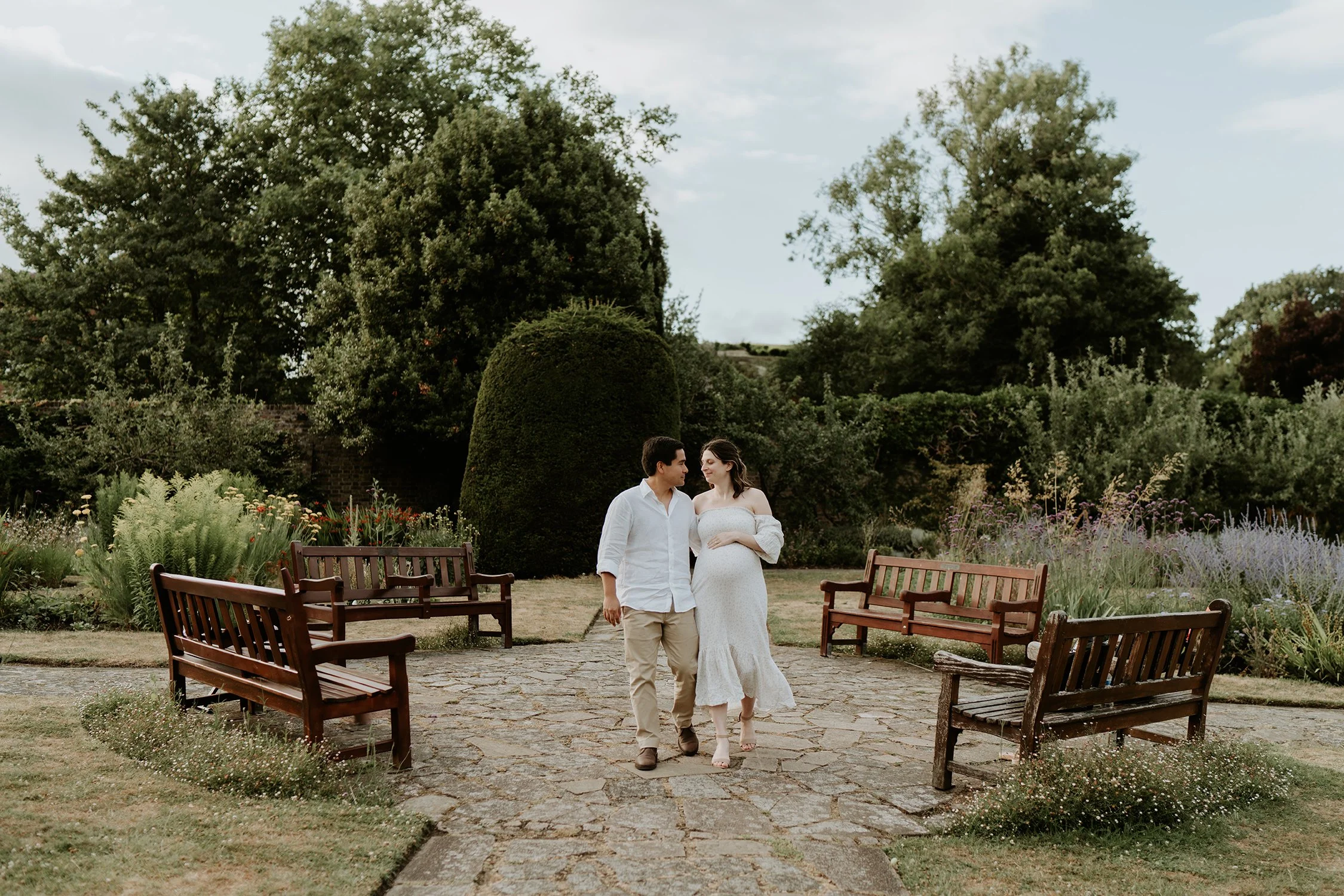 A man and a woman, with the woman being pregnant, walking on a stone path in a garden with trees and bushes, and empty wooden benches on either side.