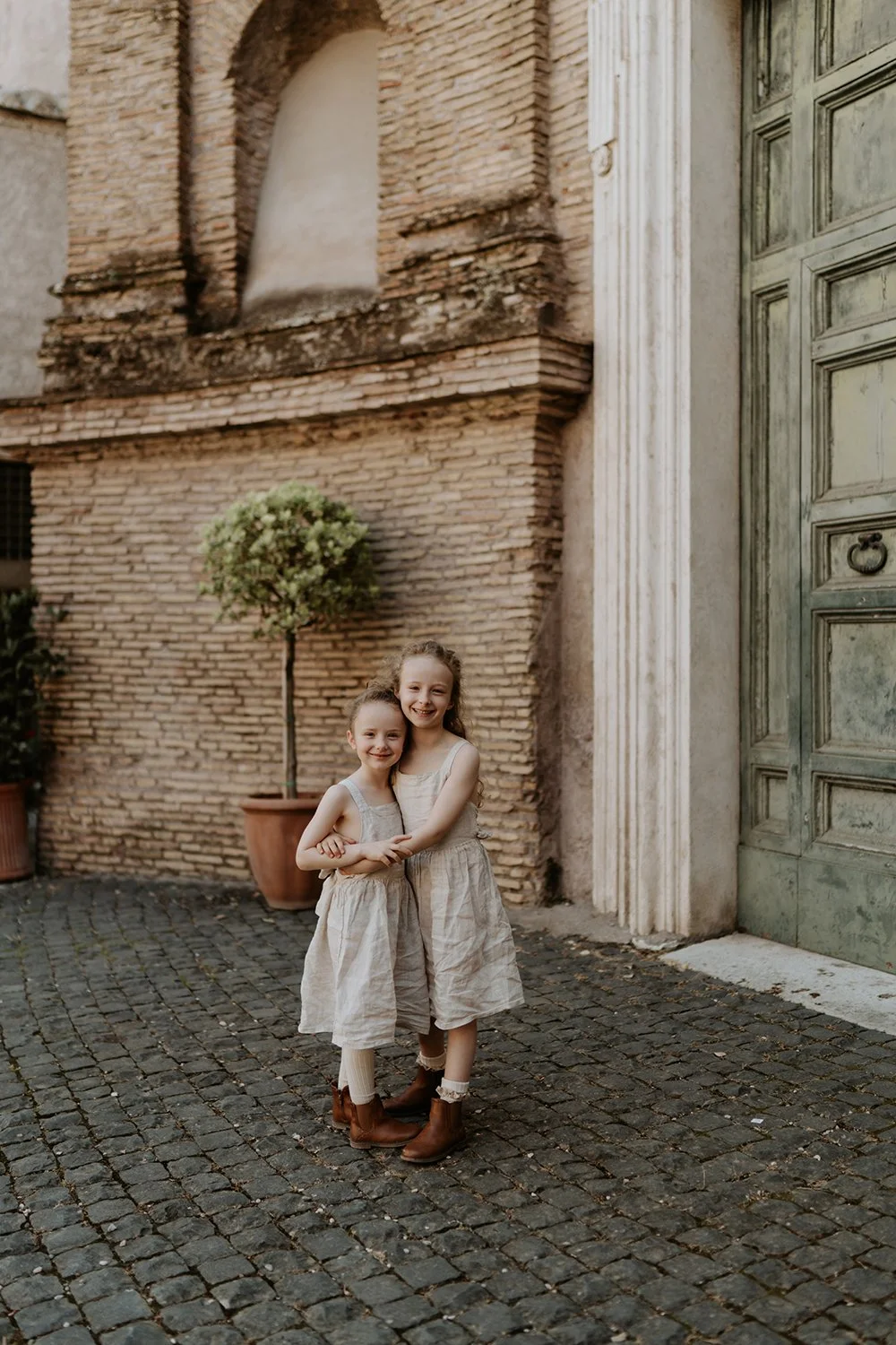 Two young girls in beige dresses and brown boots hugging and smiling on a cobblestone street in front of a brick building with a tree in a pot.