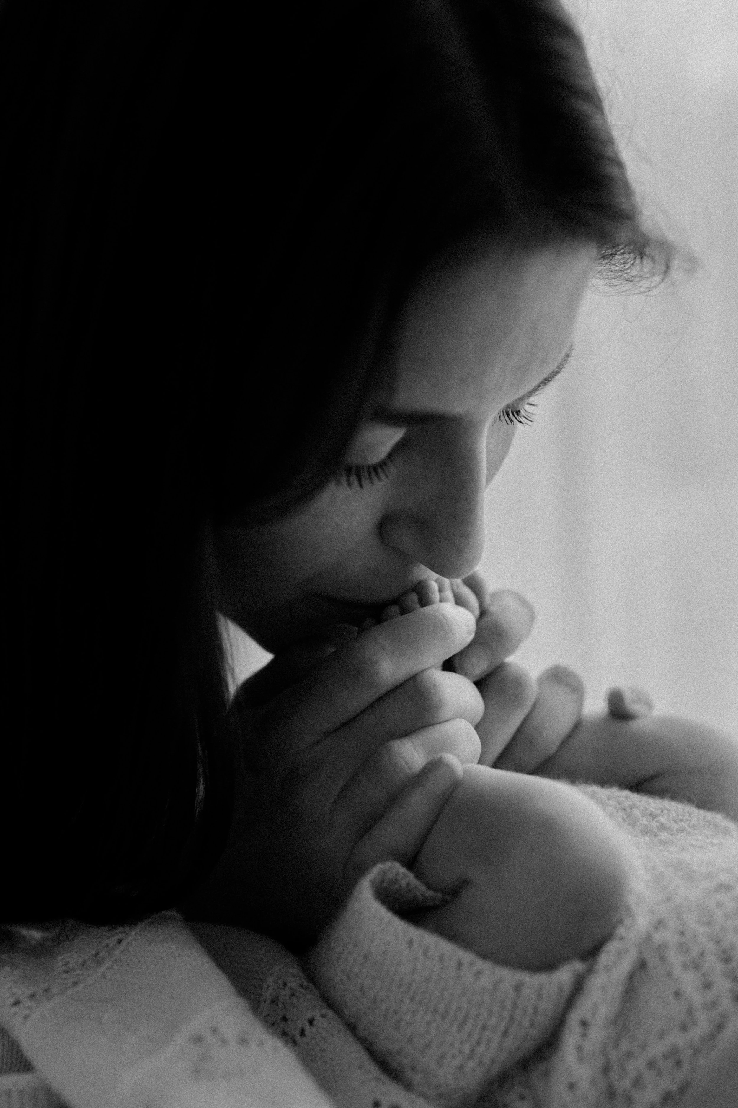 A black and white photo of a woman with her eyes closed, gently kissing or smelling a baby's foot.