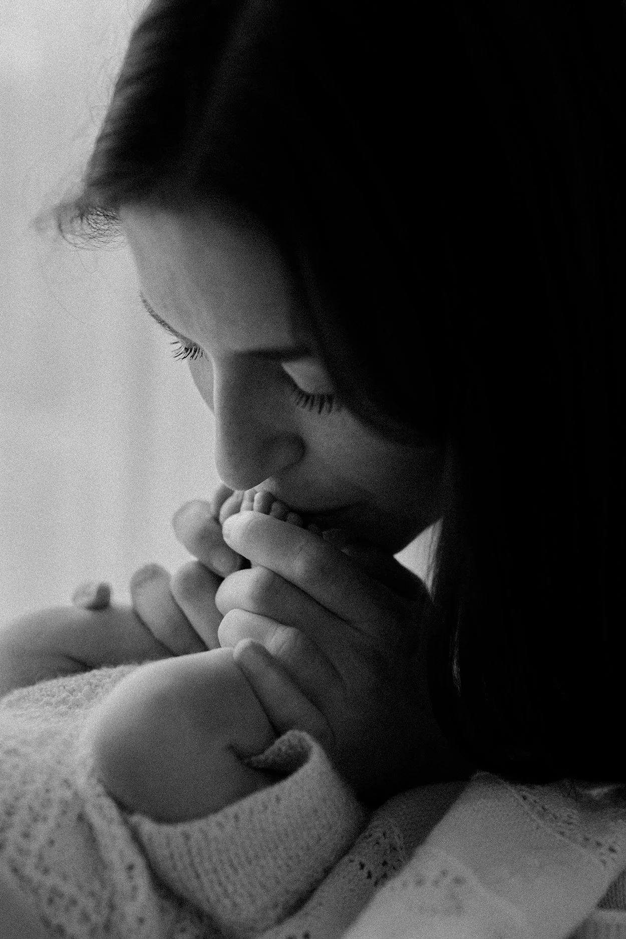 A black and white photo of a woman with closed eyes, gently kissing a baby's foot.