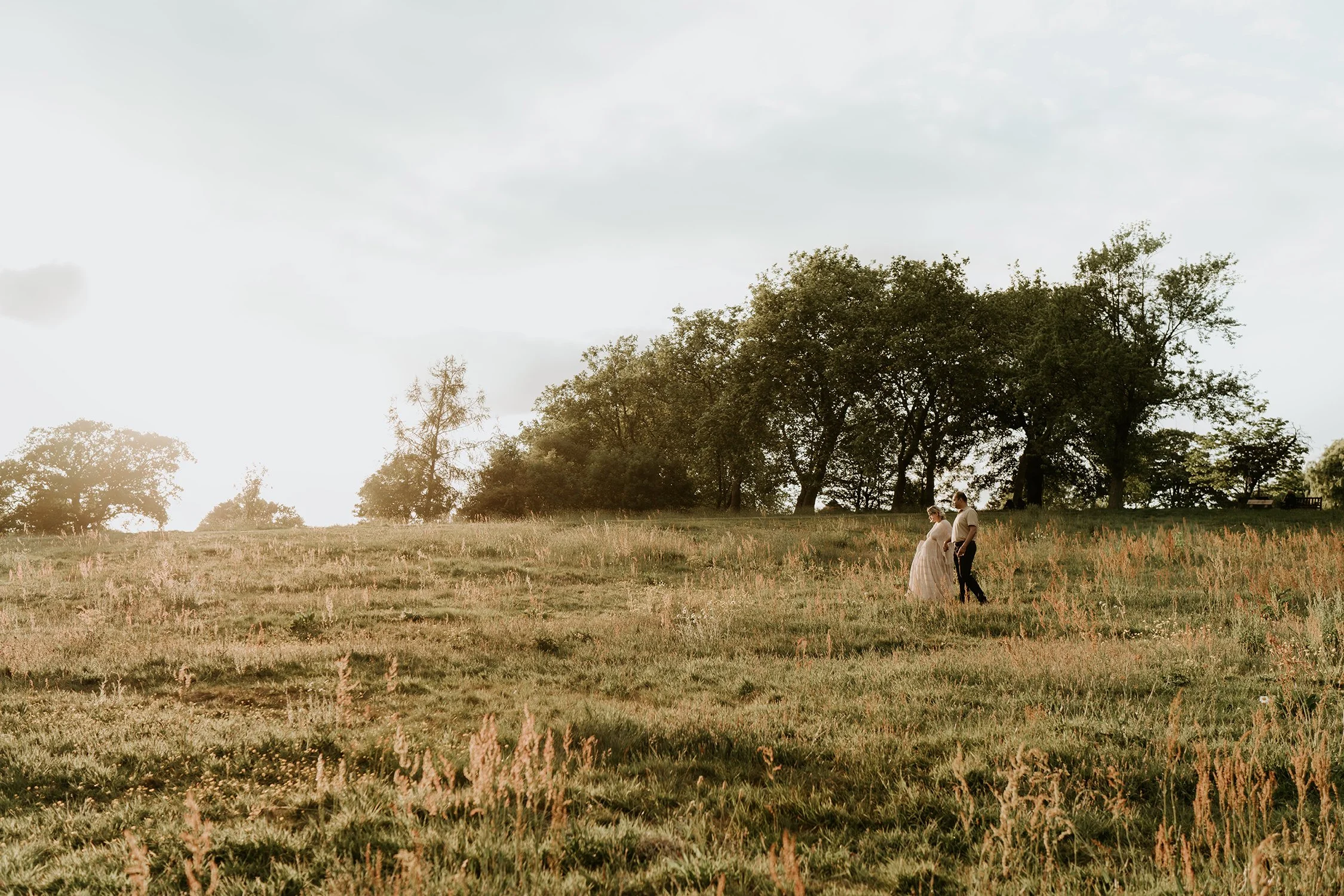 A couple holding hands stands in a grassy field with trees in the background, during sunset.