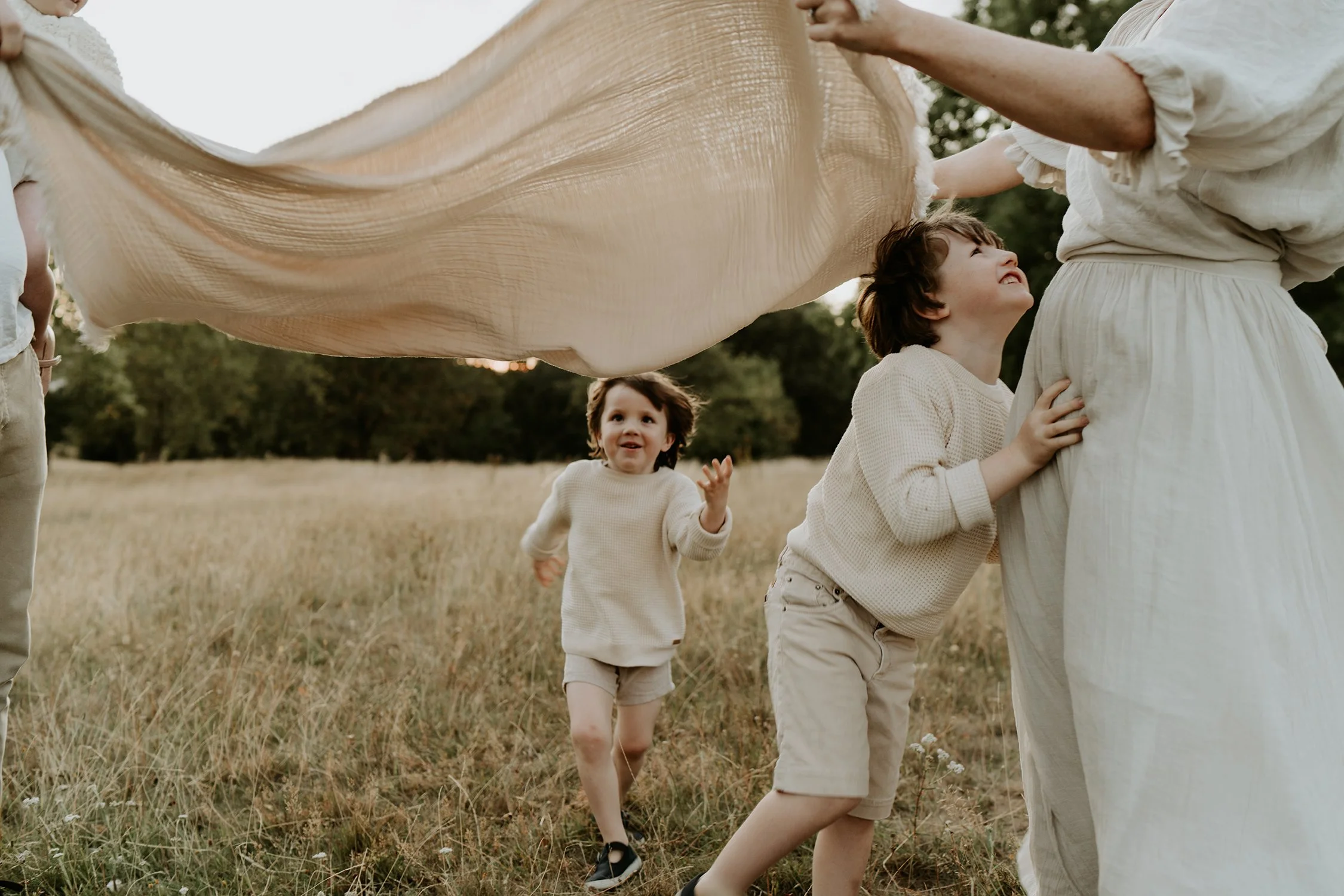 Children and an adult in casual cream-colored clothing playing outdoors with a large beige fabric, running through a grassy field.