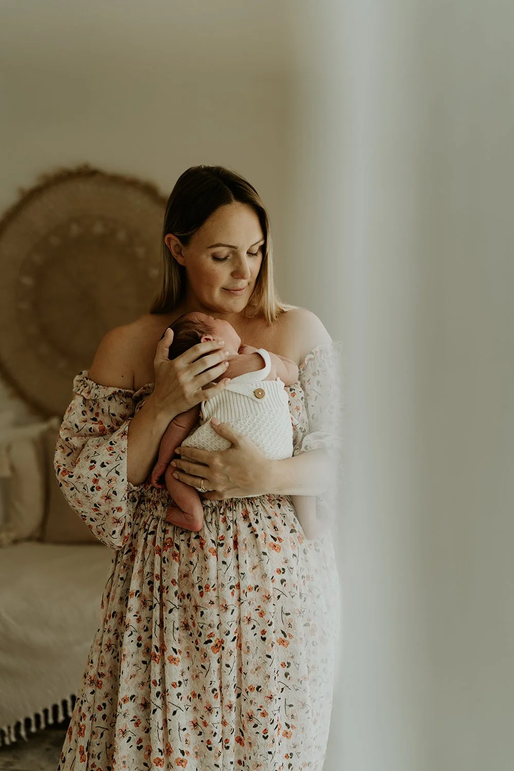 A woman with shoulder-length hair holding a newborn baby wrapped in a white blanket, standing by a window with soft light.