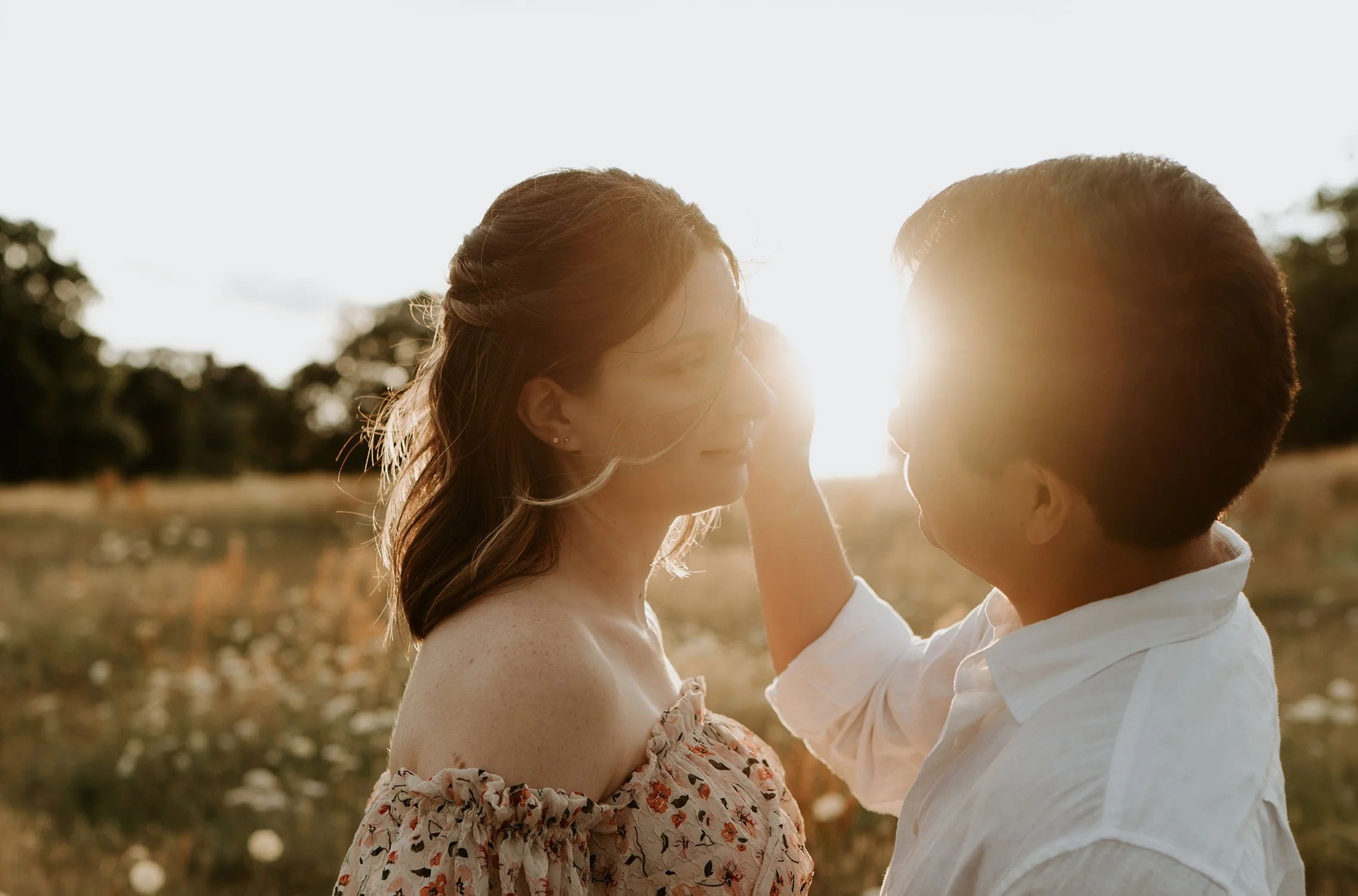 A couple is standing close to each other outdoors during sunset, with the sun behind them. The man is gently touching the woman’s face, and they are gazing into each other's eyes. The woman has long, wavy hair and is wearing a floral off-the-shoulder