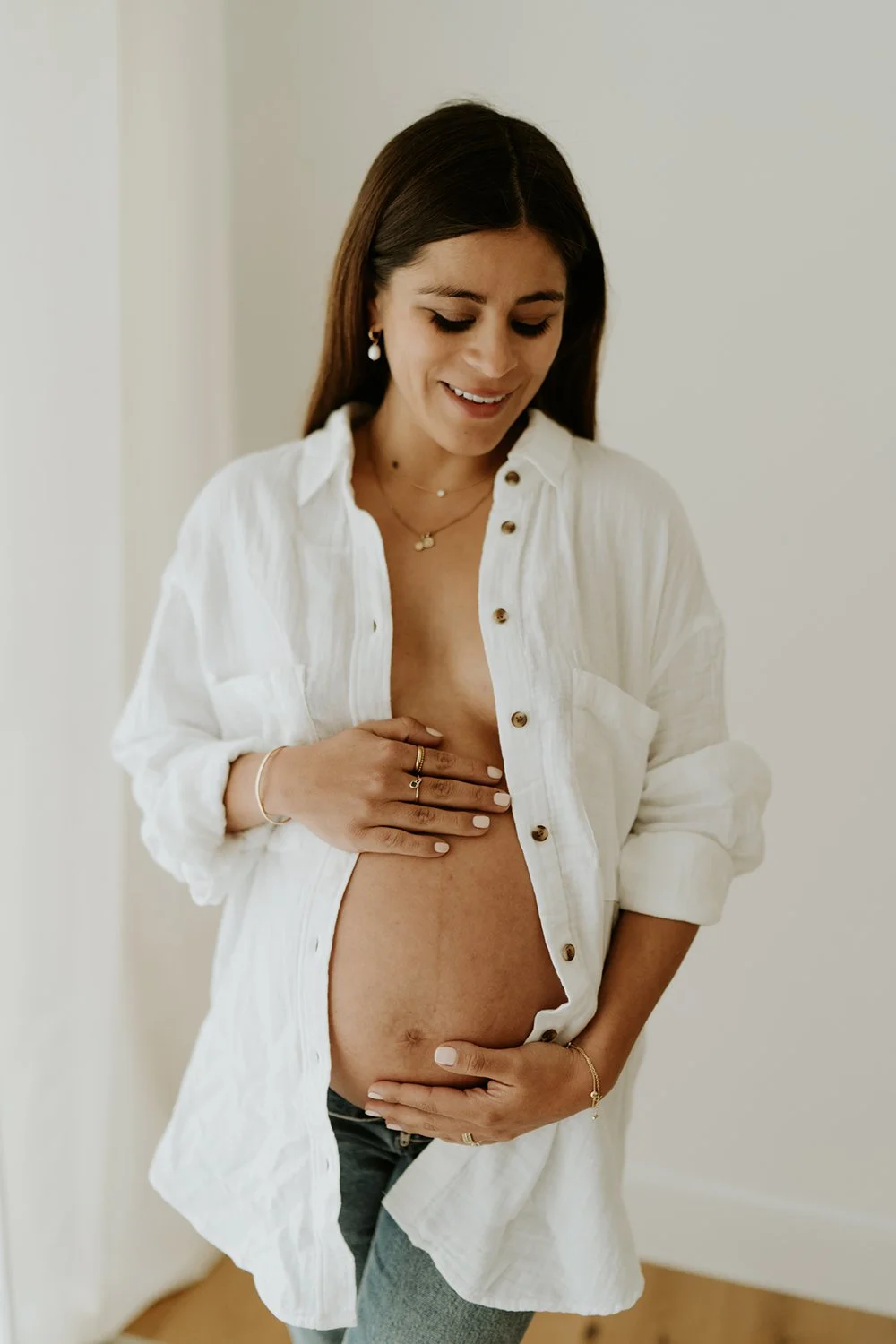 A pregnant woman with long dark hair looking down and smiling, wearing a white button-up shirt partially open, revealing her pregnant belly, accessorized with jewelry.