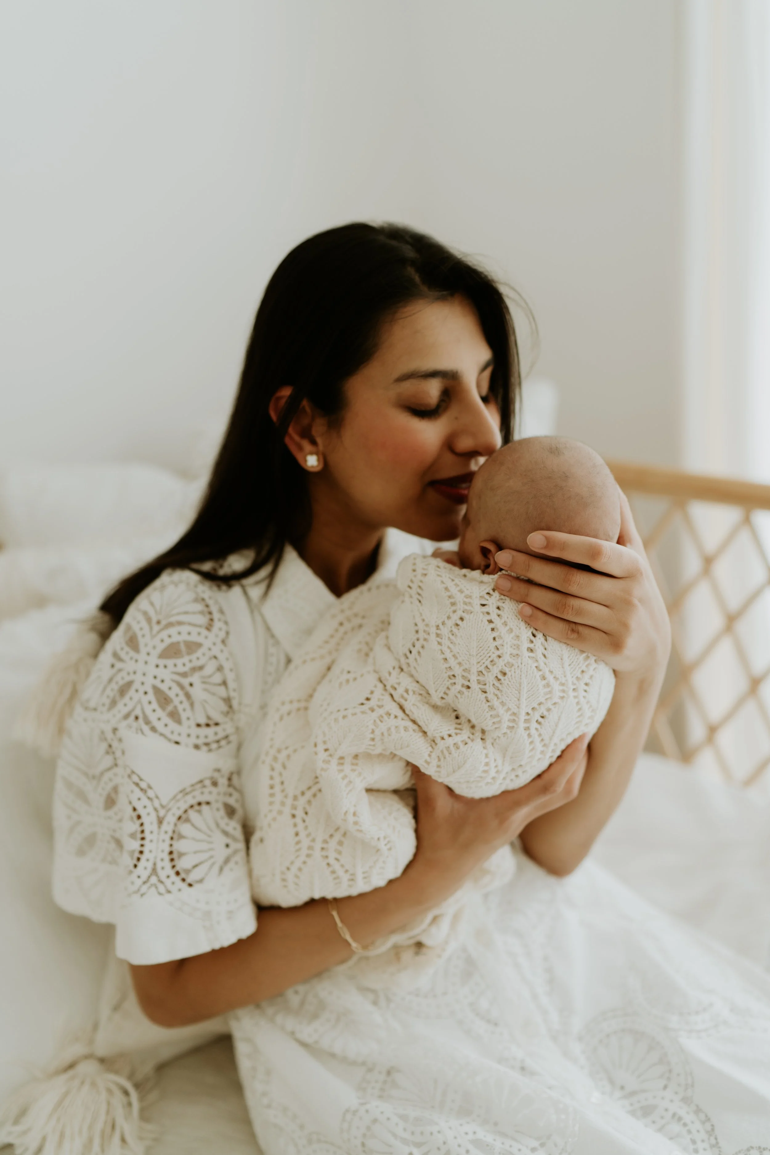 A woman cuddling and kissing a baby wrapped in a cream-colored, crochet blanket.