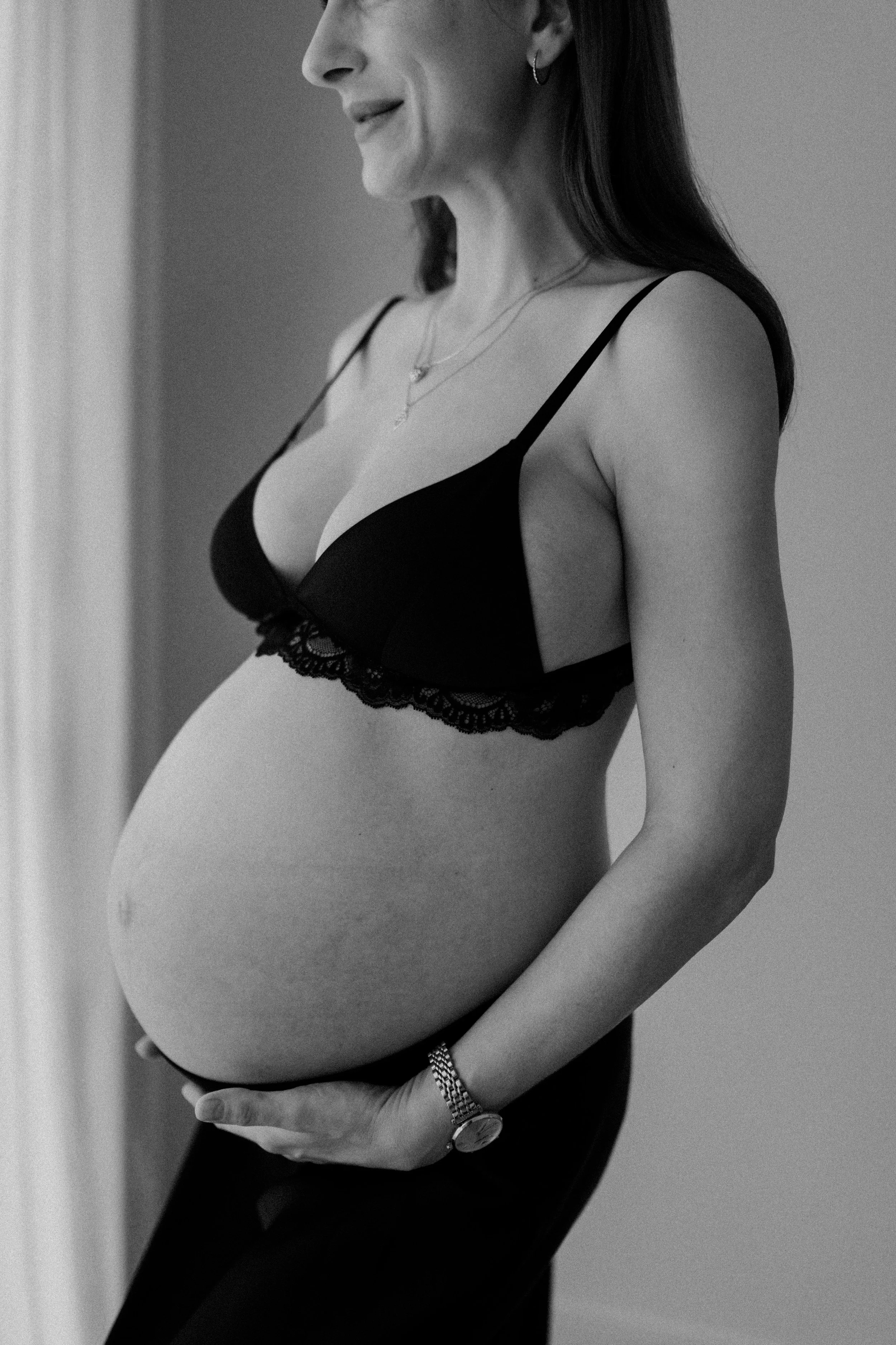 A pregnant woman wearing black lingerie, showing her pregnant belly, with her hand resting on her stomach, and jewelry including earrings, necklaces, and a watch, in black and white.