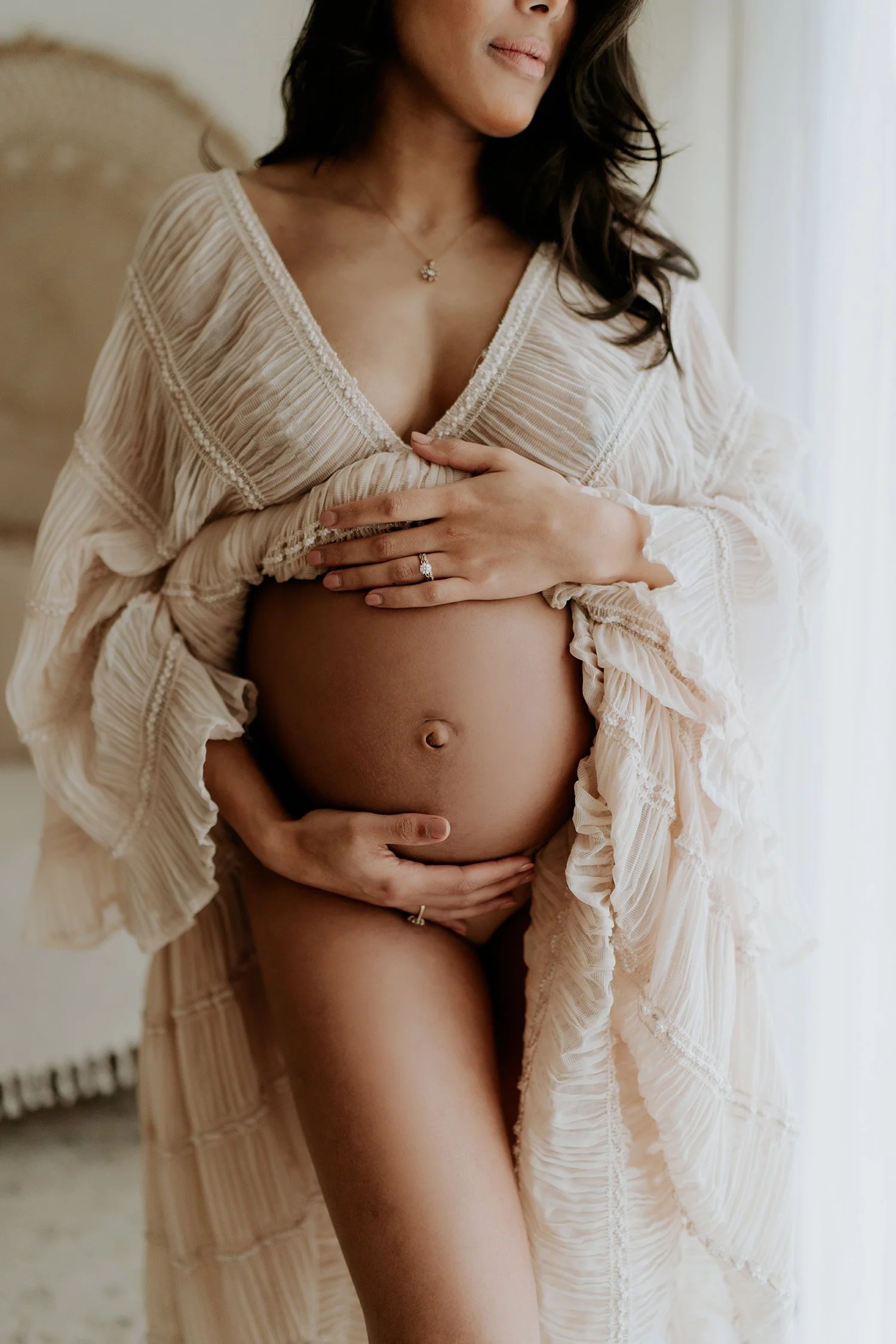 A pregnant woman holding her belly with both hands, wearing a beige dress with lace details, standing indoors near a window with white curtains.