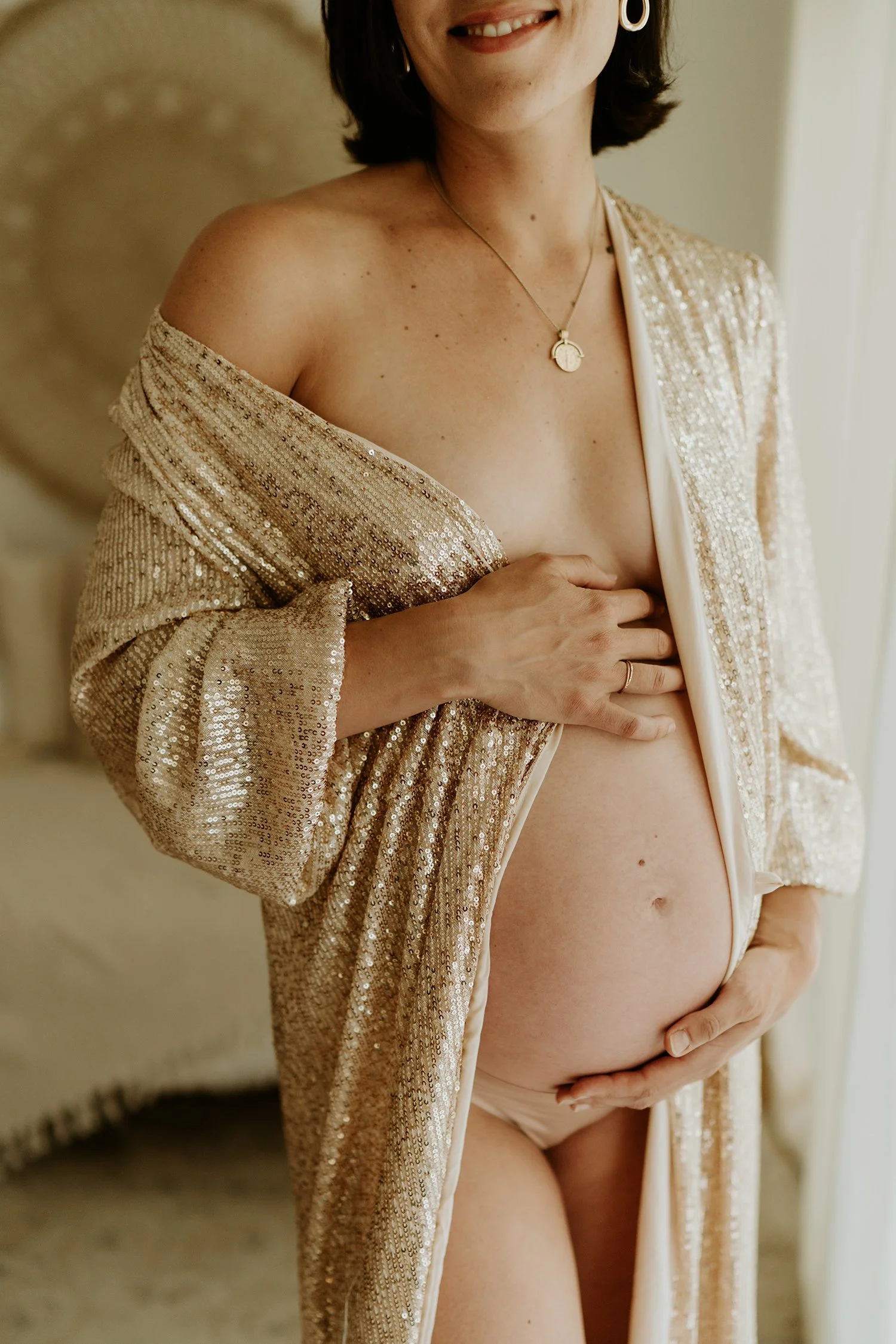 A pregnant woman in a gold, sequined robe partially open, cradling her belly, standing near a window with natural light.