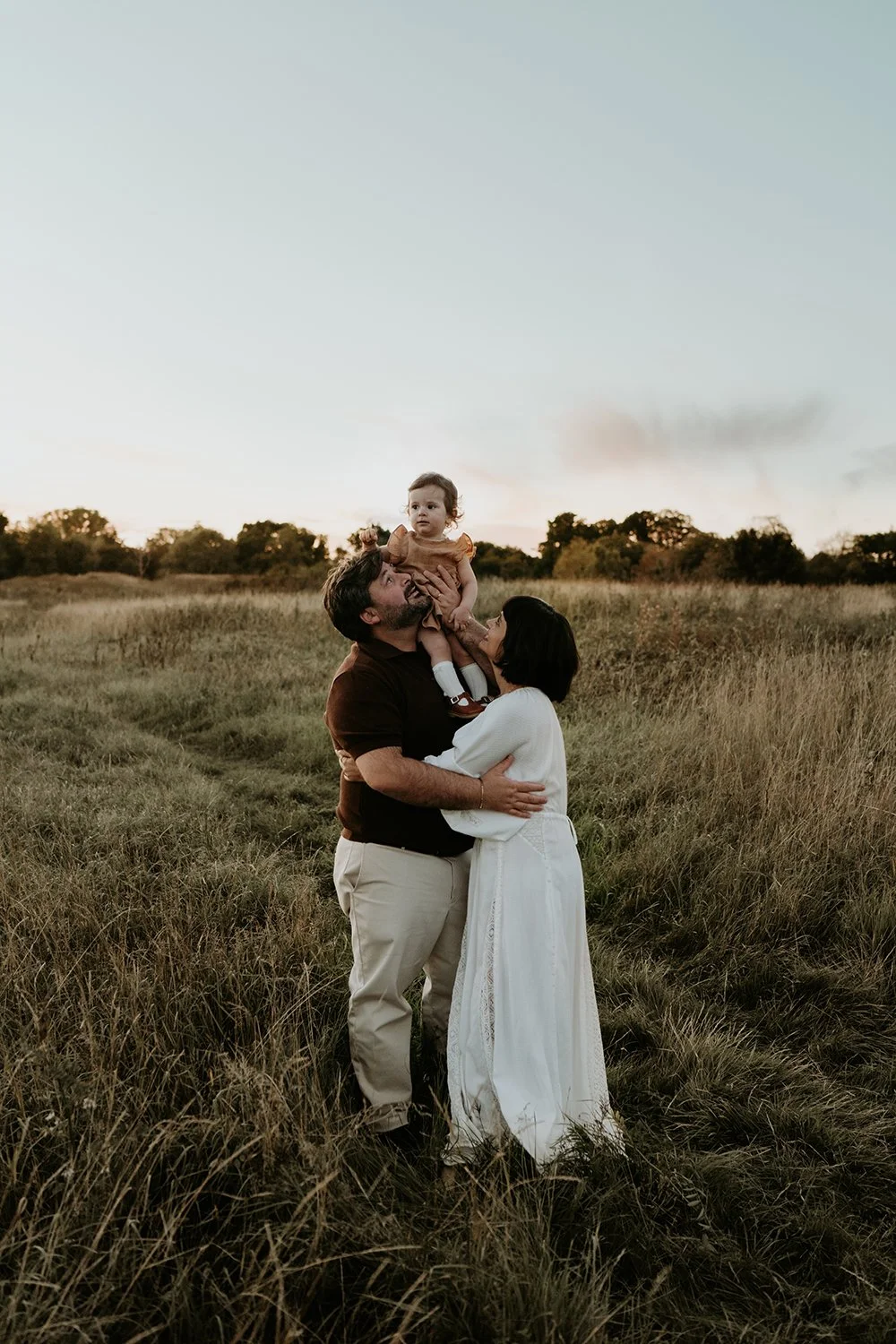 A family of three in a grassy field during sunset, with the father lifting a young girl in the air, and the mother standing close, all smiling and enjoying the moment.
