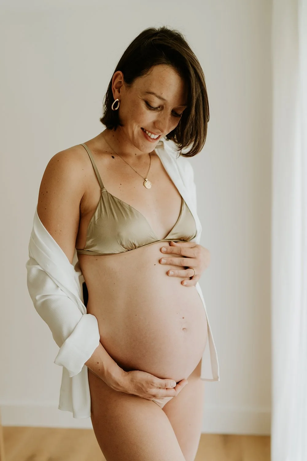 A pregnant woman smiling, wearing a beige bra and white shirt draped over her shoulders, standing in a softly lit room with a neutral background.