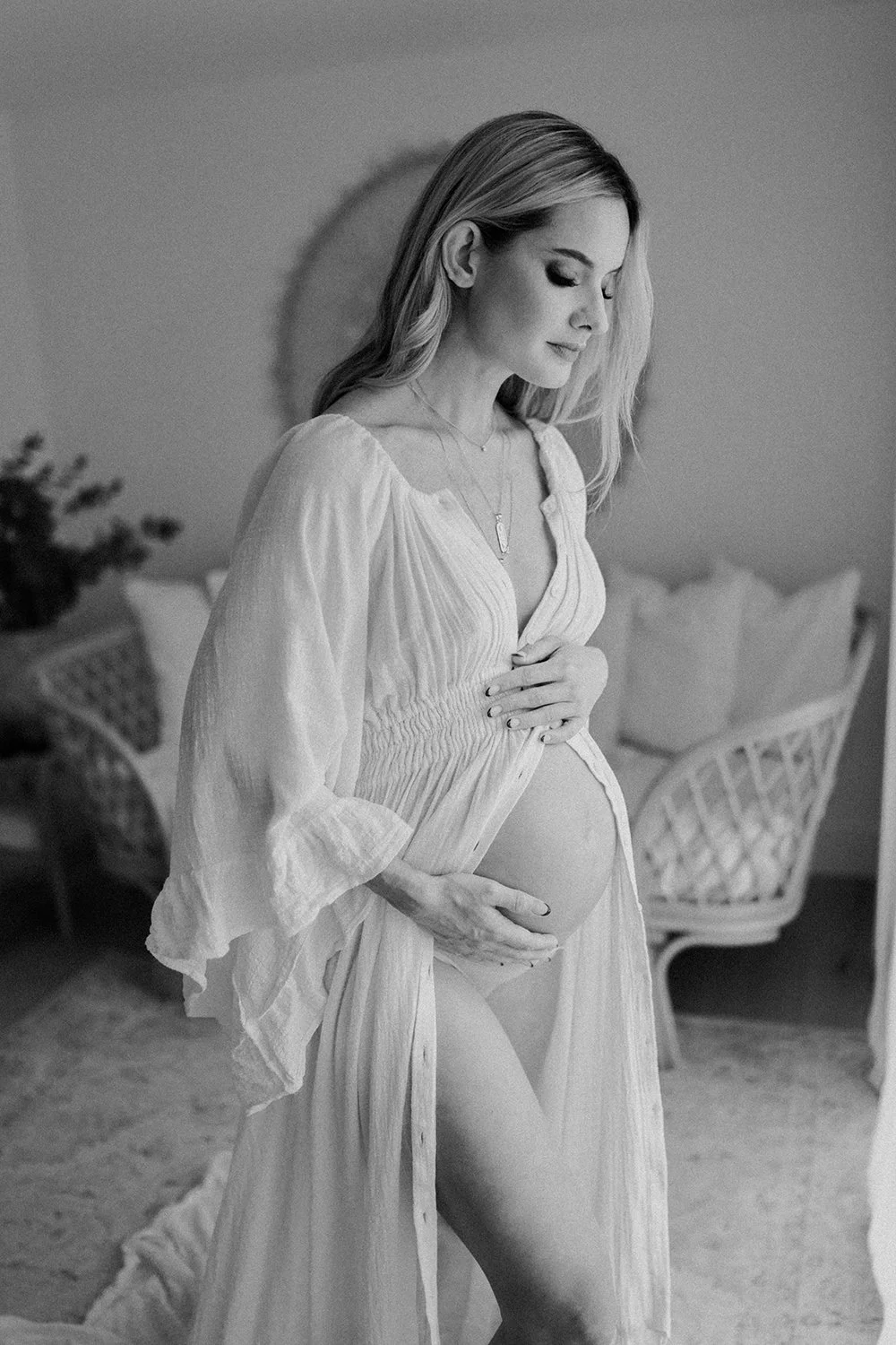Black and white photograph of a pregnant woman wearing a light, flowing dress, gently holding her belly with one hand. She has long, wavy hair and a serene expression, standing in a room with wicker furniture and soft pillows.