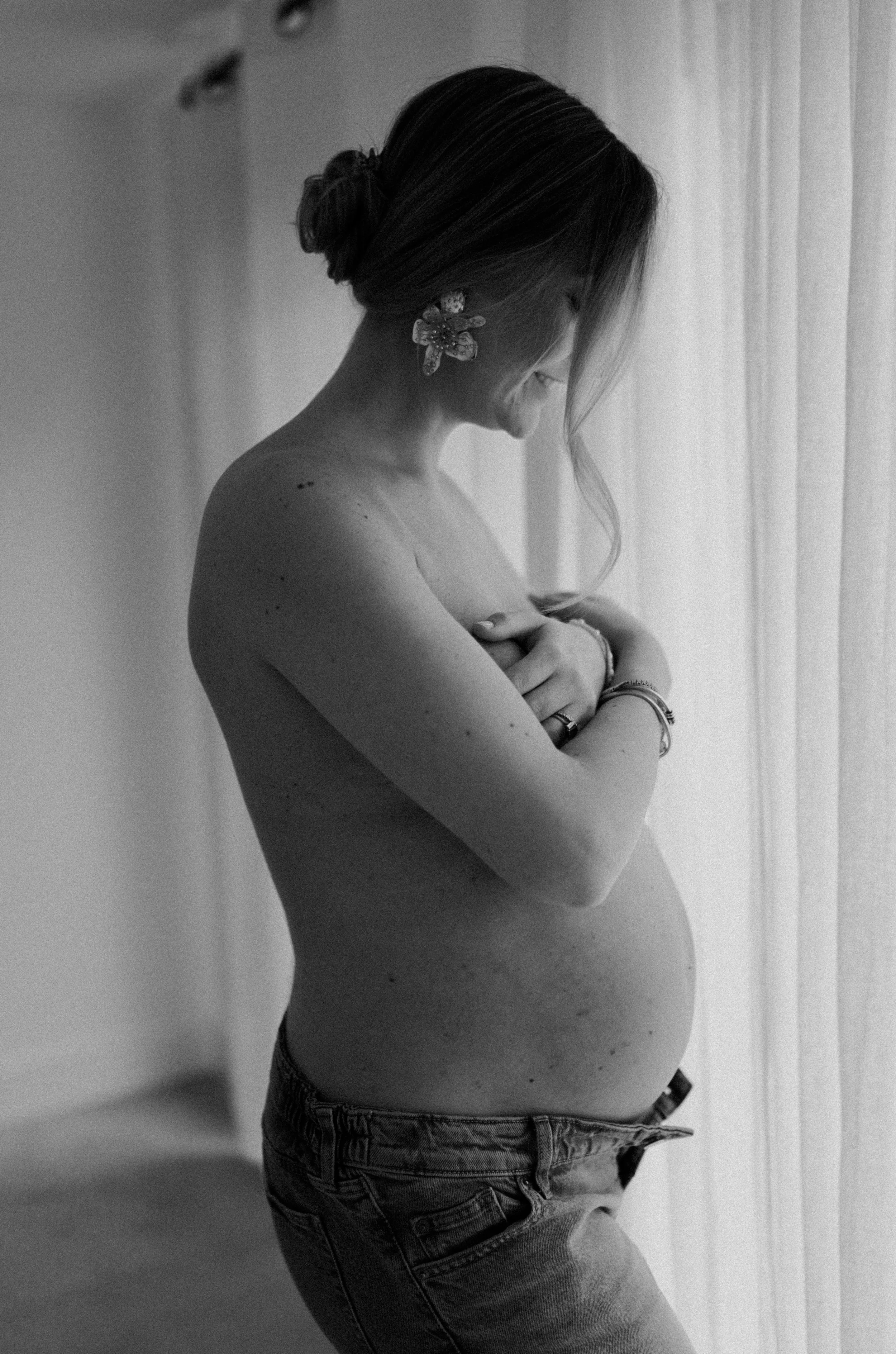 A pregnant woman standing sideways, with her eyes closed, arms crossed over her chest, wearing a flower-shaped earring and jeans, near a window with curtains, in black and white.