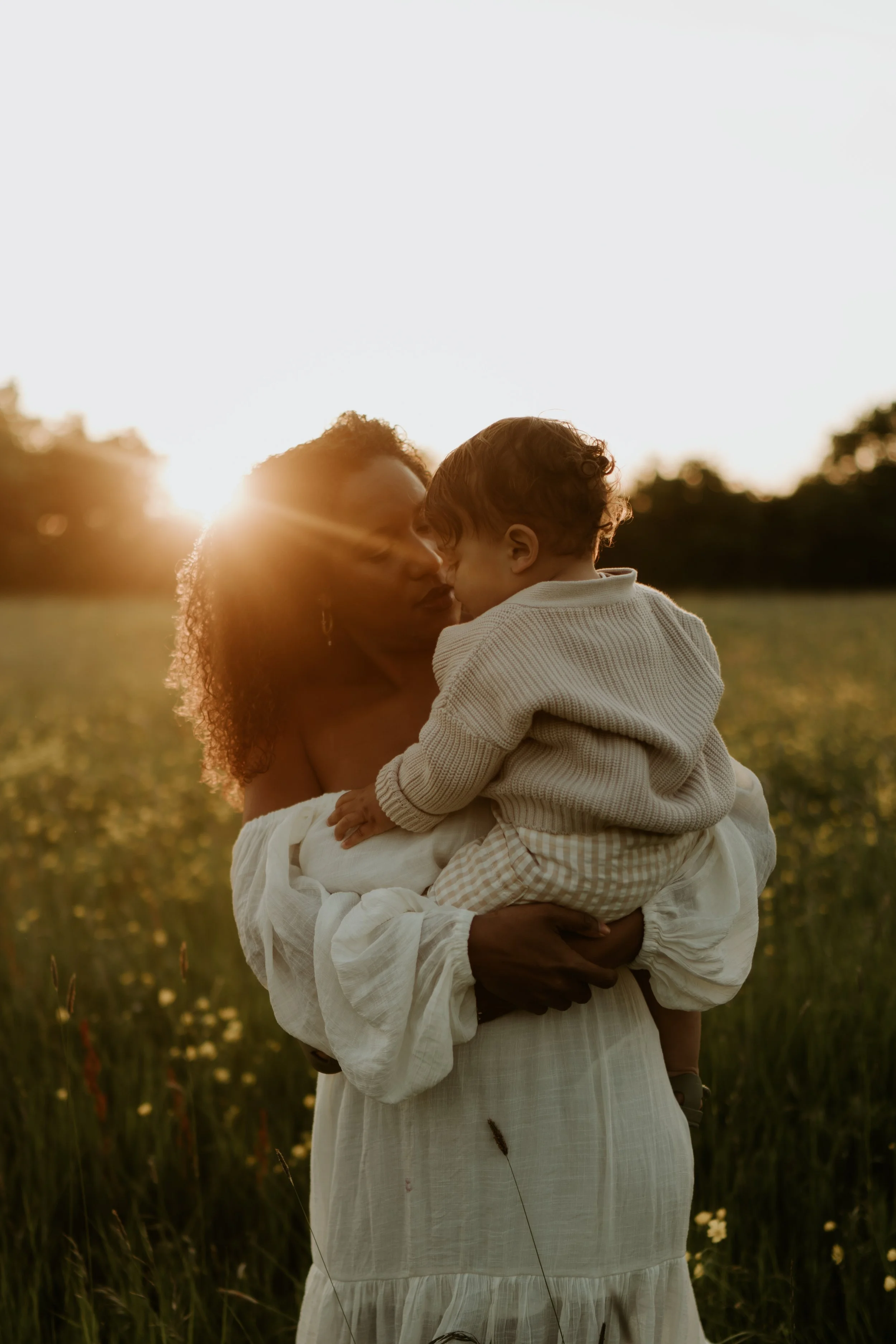 A woman holding a young child in a field during sunset, with the sun shining behind her.