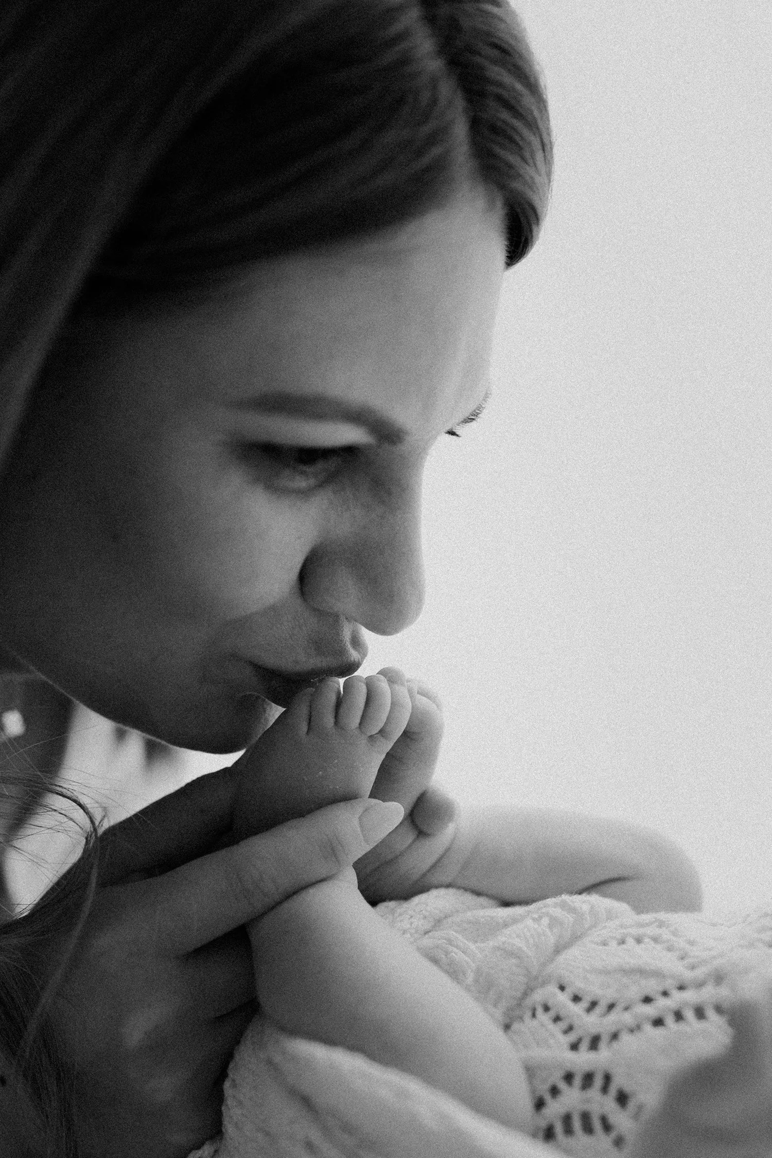 Close-up of a woman with closed eyes and long hair whispering into a baby's ear, with hands gently holding the baby. The scene is in black and white.
