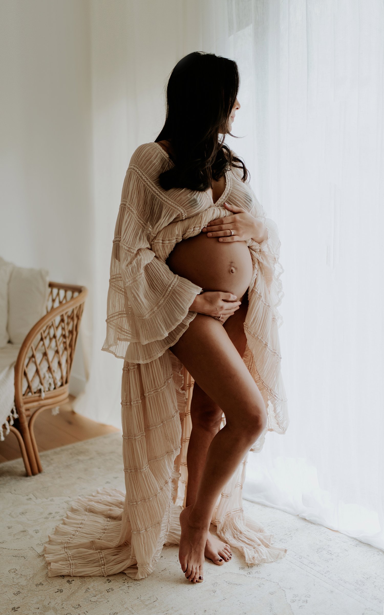 A pregnant woman stands indoors near a window with sheer curtains, wearing a light-colored, flowing, pleated dress. She gently holds her belly with one hand and looks outside, with her face in profile. The room has a wicker chair and a beige patterne