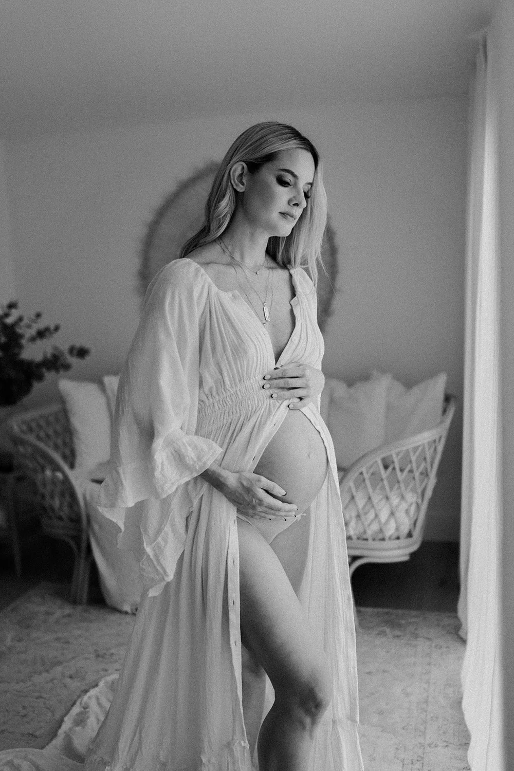 Black and white photo of a pregnant woman in a flowing dress, standing in a room with wicker furniture and a plant, gently touching her belly and looking down.